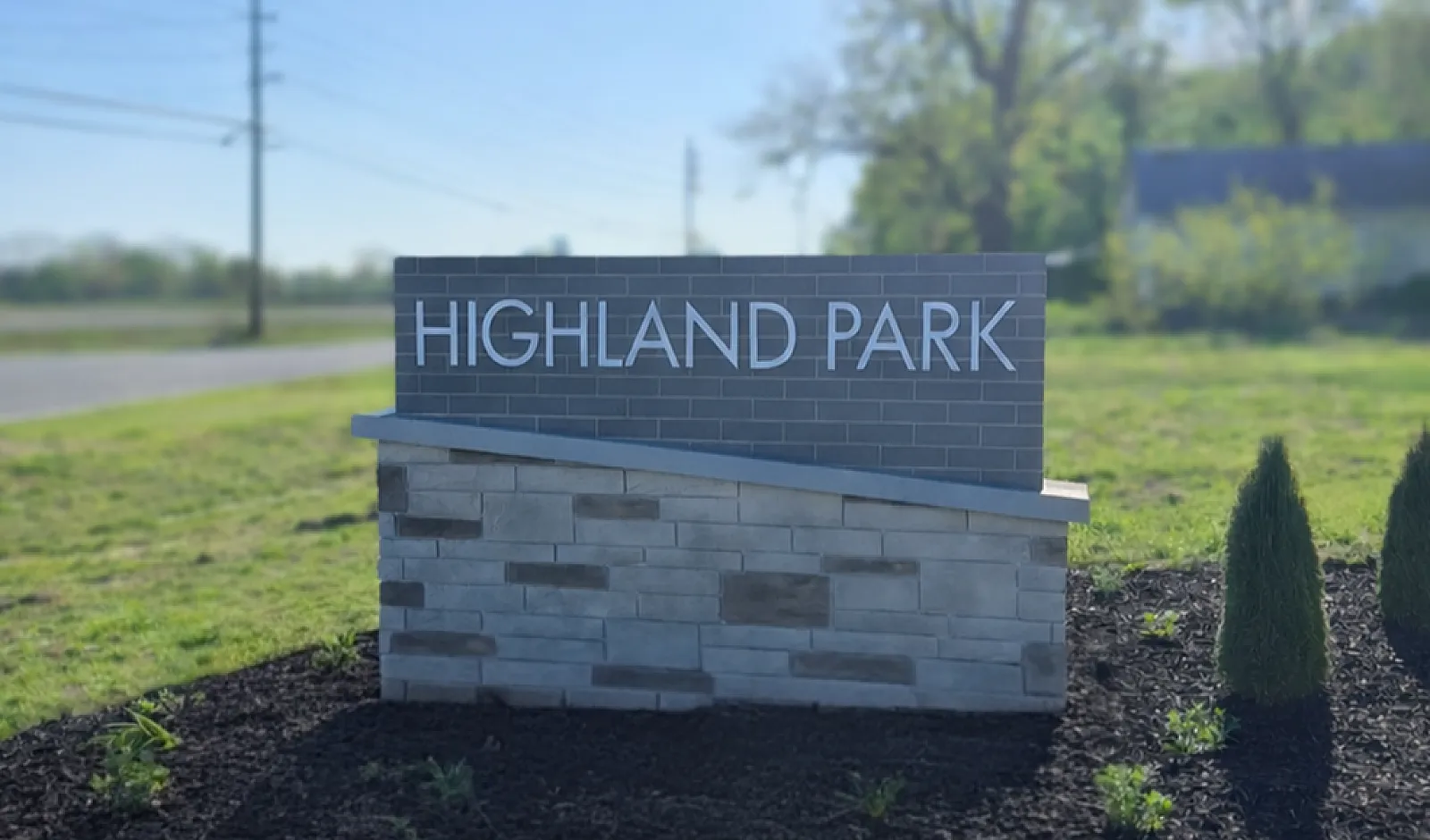 Highland Park entrance sign with a modern stone base and lush greenery under a bright, sunny sky.