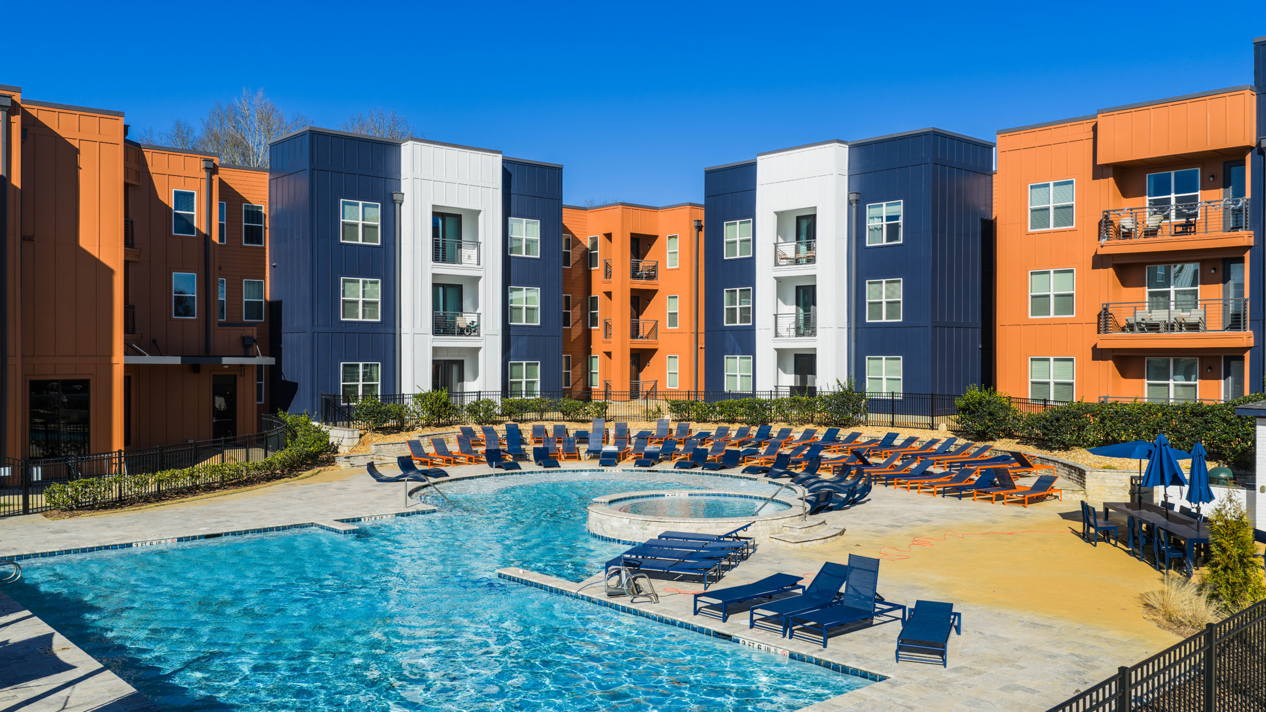 Vibrant modern apartment complex with spacious pool area, surrounded by sun loungers and colorful buildings under a clear blue sky.