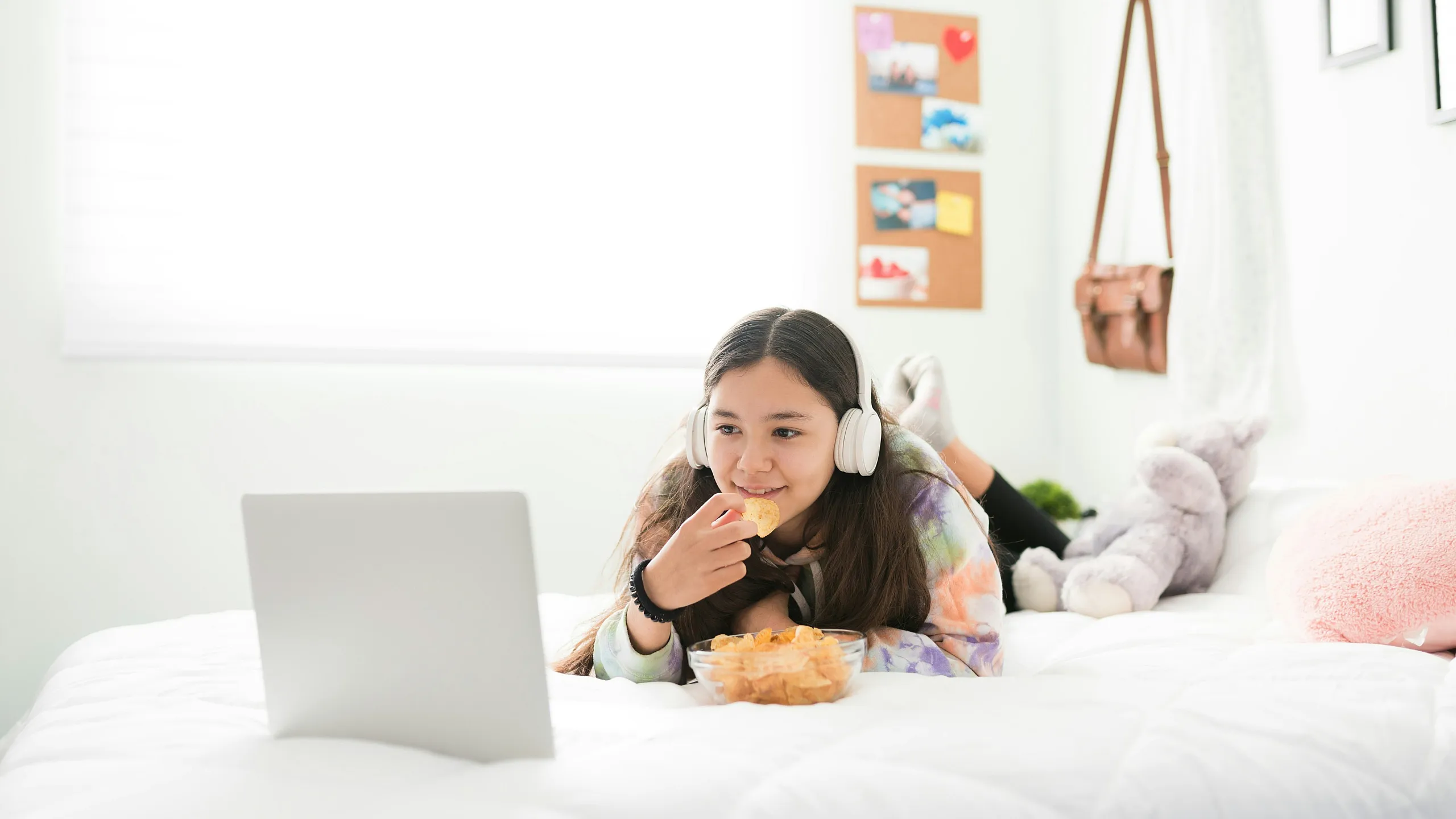Cozy bedroom scene with a young girl enjoying snacks on a white bed, wearing headphones and using a laptop, surrounded by plush toys and colorful decor.