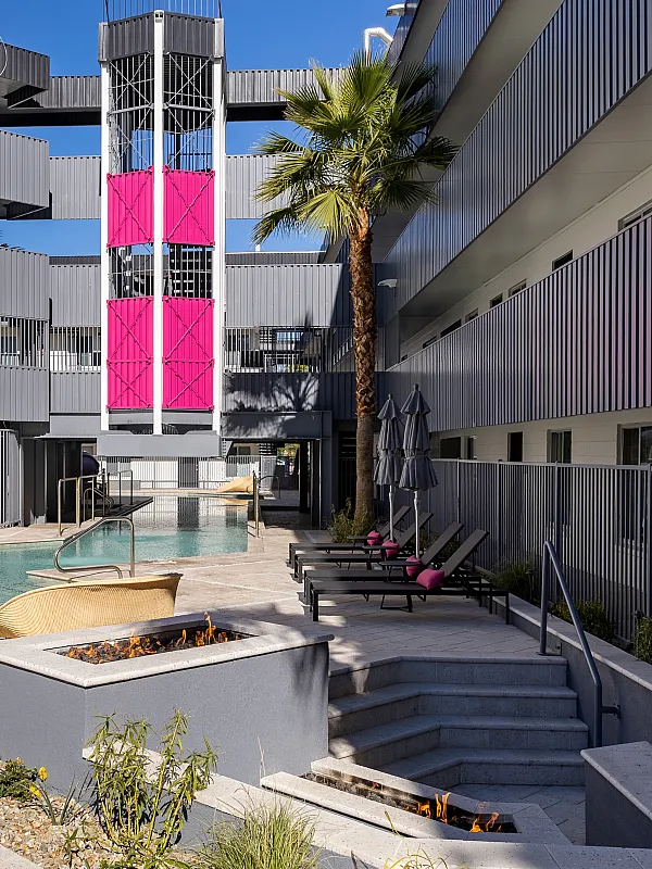Modern apartment courtyard featuring a sleek pool, stylish lounge chairs with pink accents, and a vibrant palm tree under a clear blue sky.