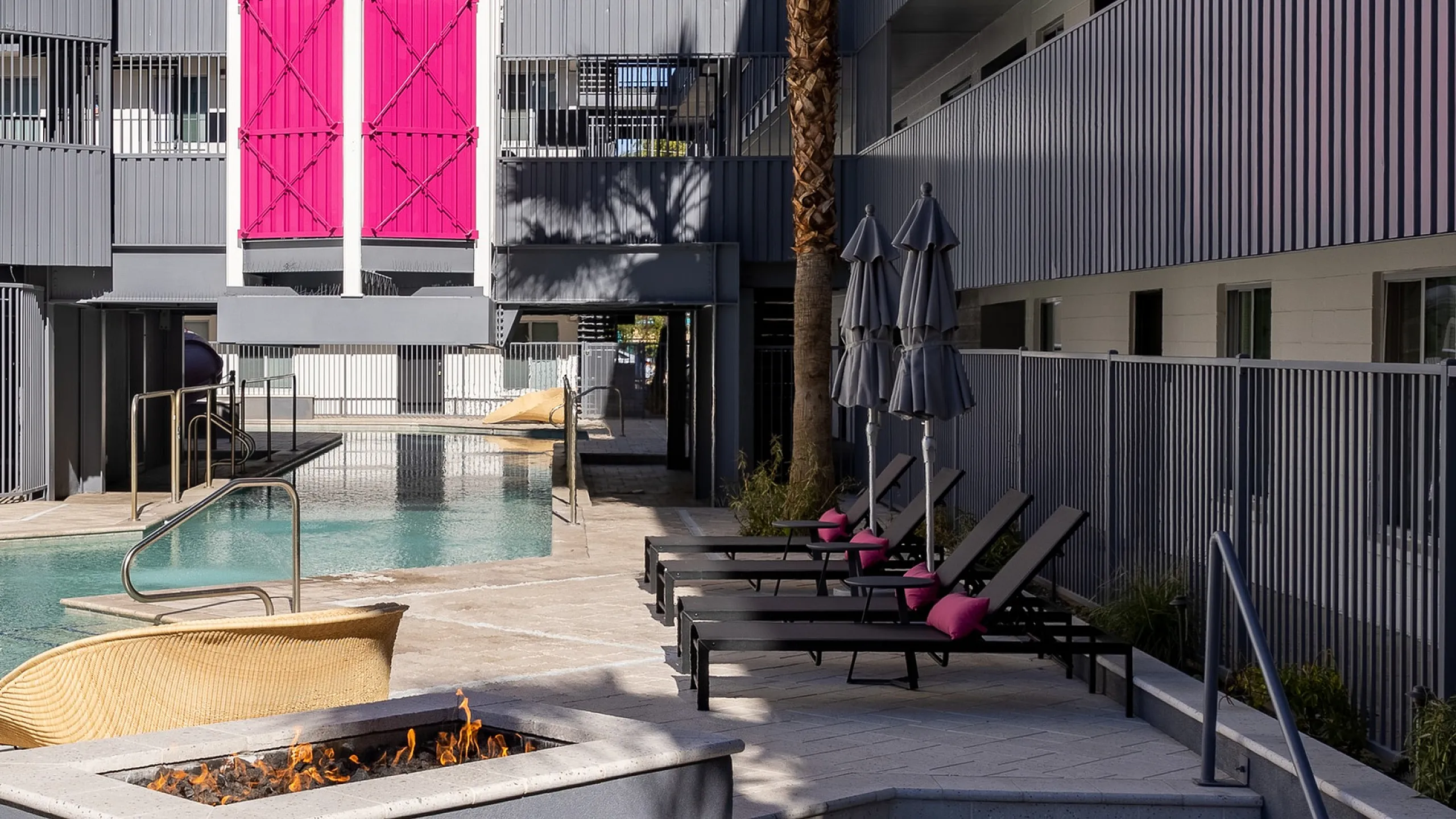 Contemporary apartment complex courtyard featuring a sleek swimming pool, chic lounge chairs with pink accents, vibrant palm tree, modern architecture, and cozy fire pit.