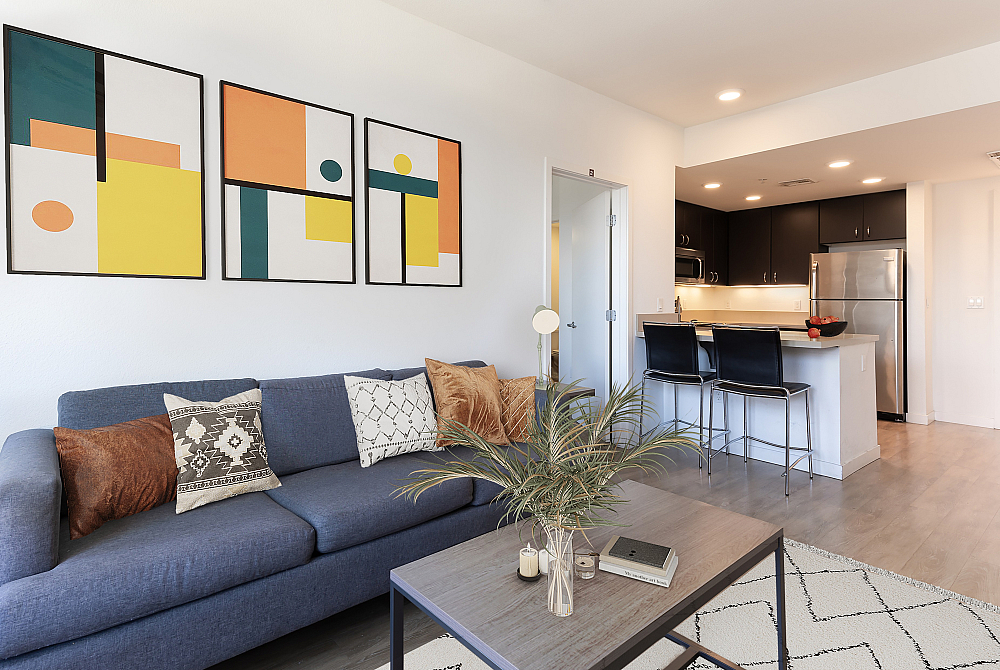 Modern living room with stylish decor and open kitchen, featuring a comfortable blue sofa, contemporary artwork, and sleek black bar stools.