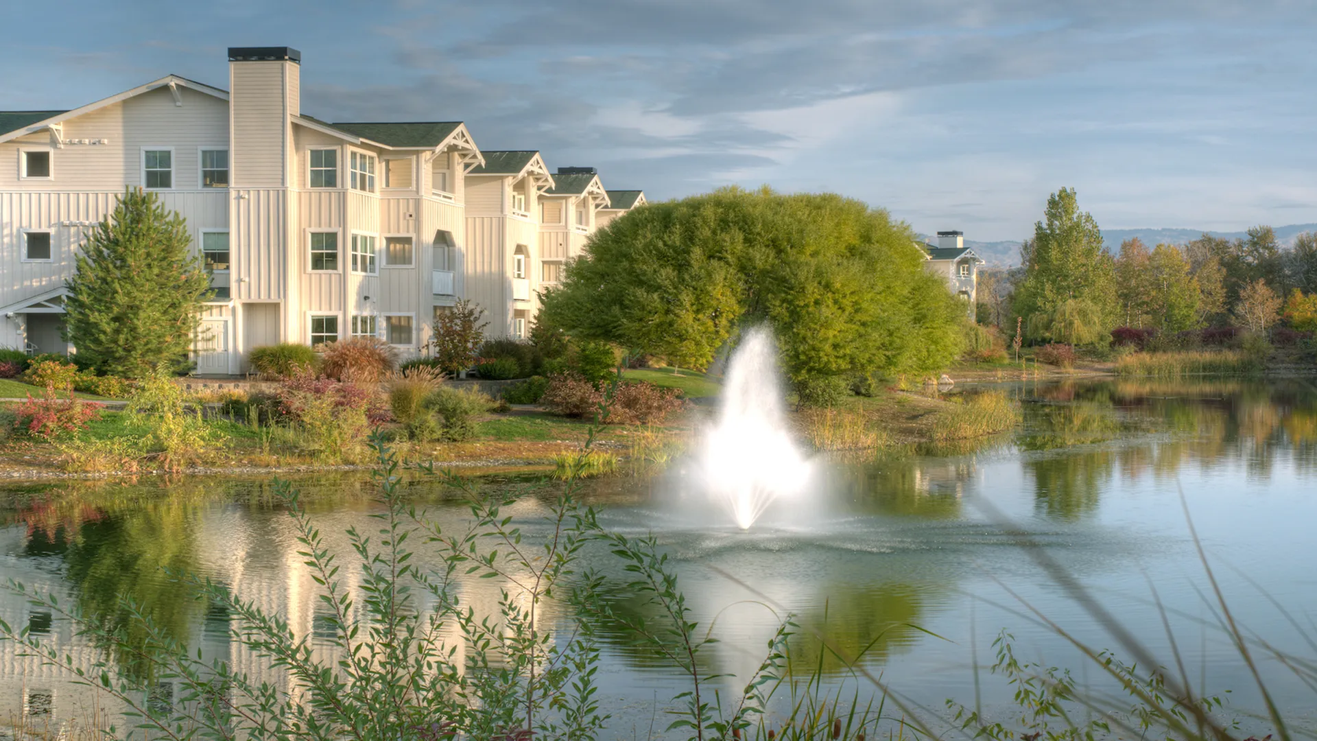 Picturesque apartment complex with modern architecture, surrounded by lush greenery and a serene pond with a striking fountain.
