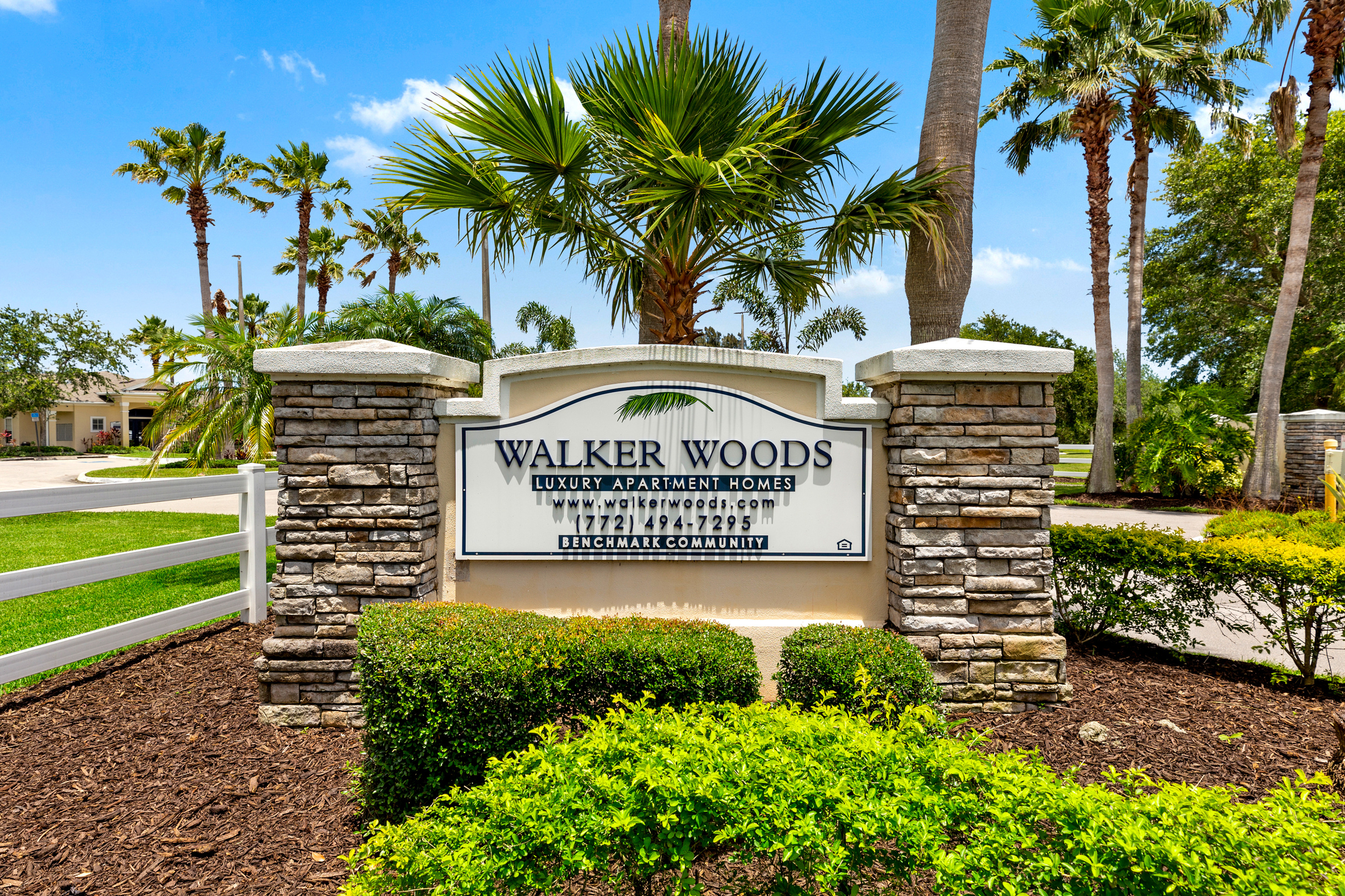 Inviting entry sign for Walker Woods Luxury Apartment Homes surrounded by lush greenery and palm trees under a clear blue sky.