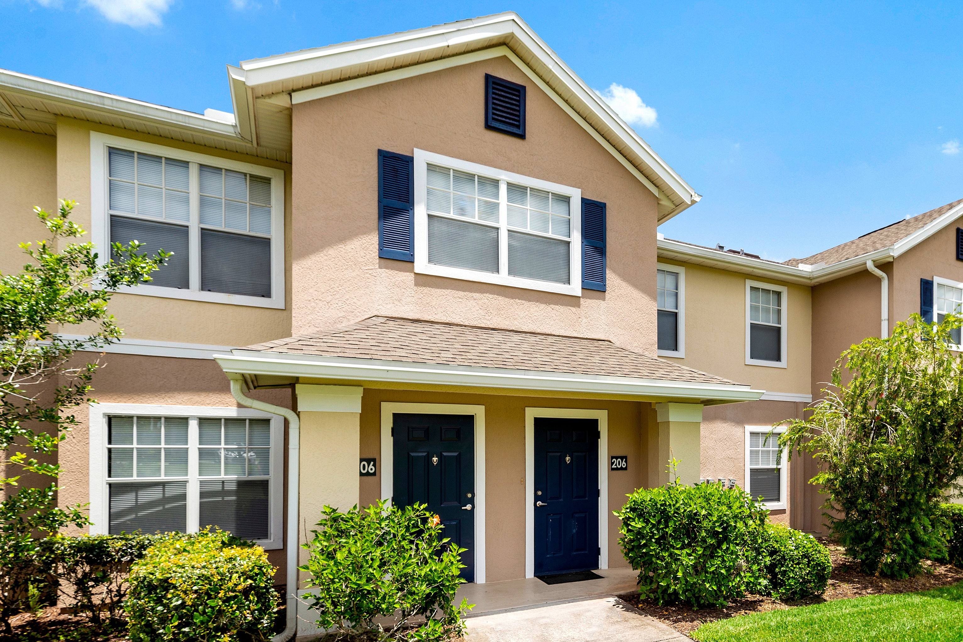 Charming townhouse exterior with dual navy front doors, surrounded by vibrant greenery and blue shutters under a clear sky.