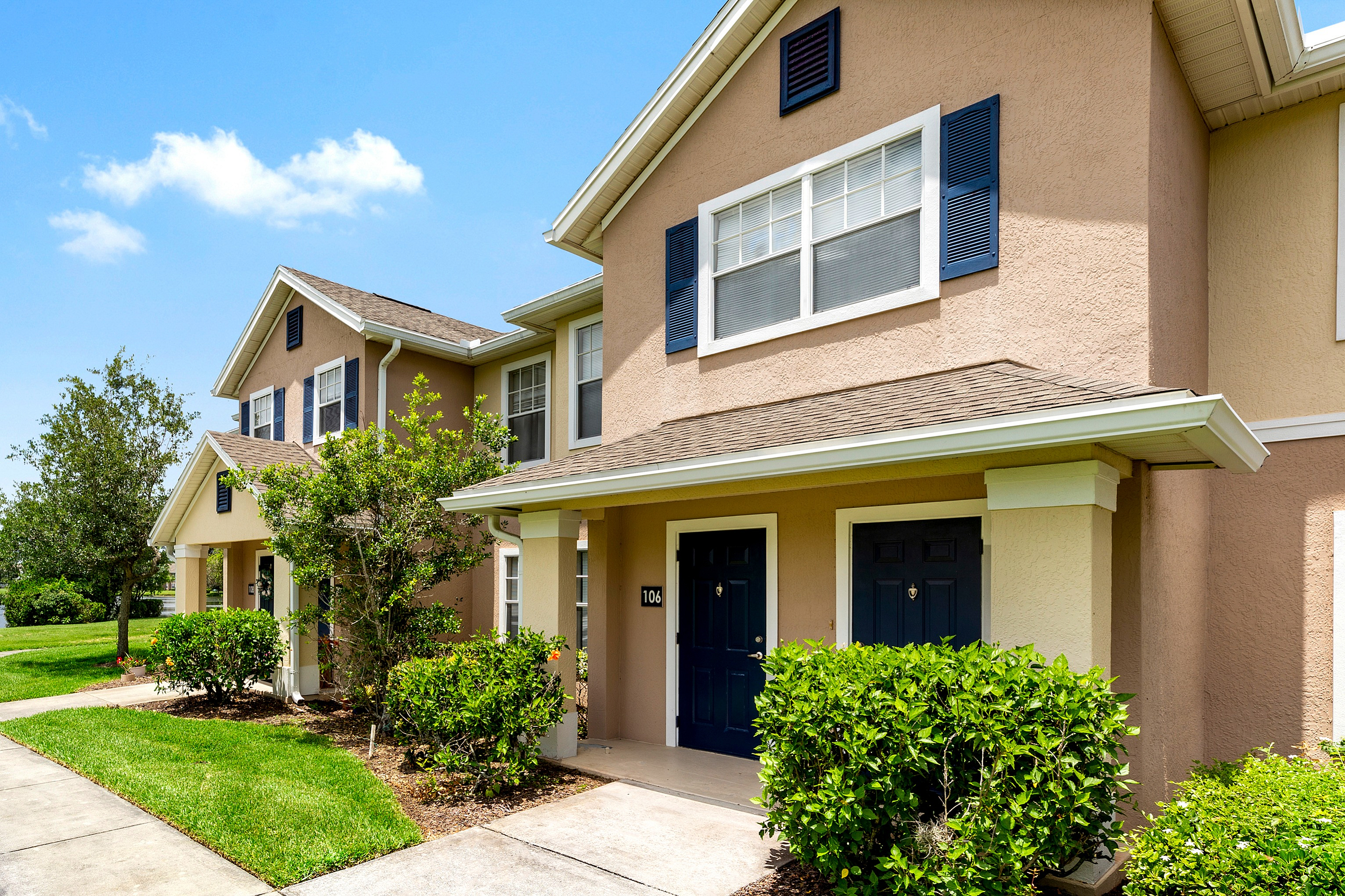 Charming two-story townhouse with well-maintained greenery, inviting front entrance, and elegant shutters, set under a clear blue sky.