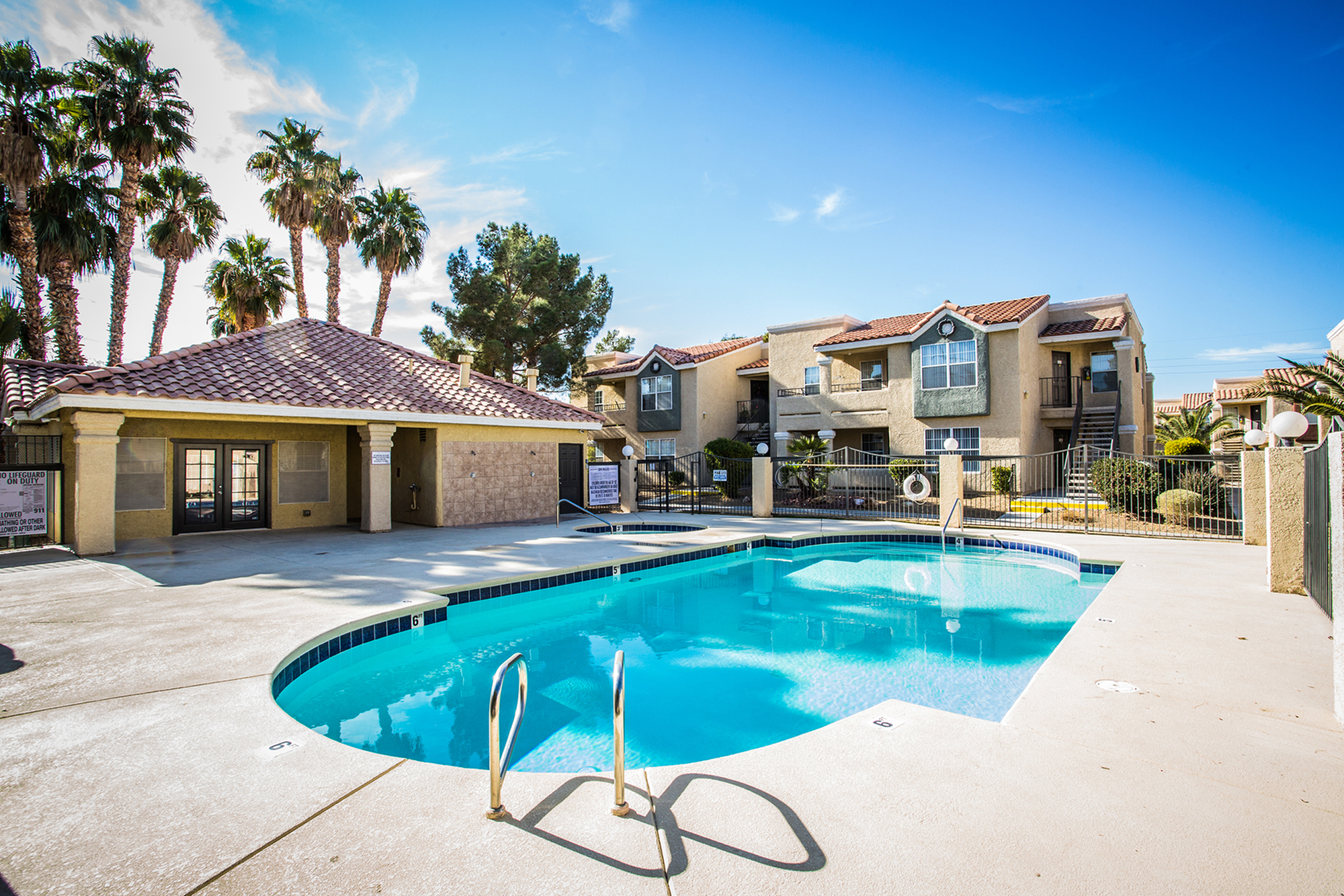 Inviting apartment complex with a sparkling swimming pool, surrounded by lush palm trees under a clear blue sky.