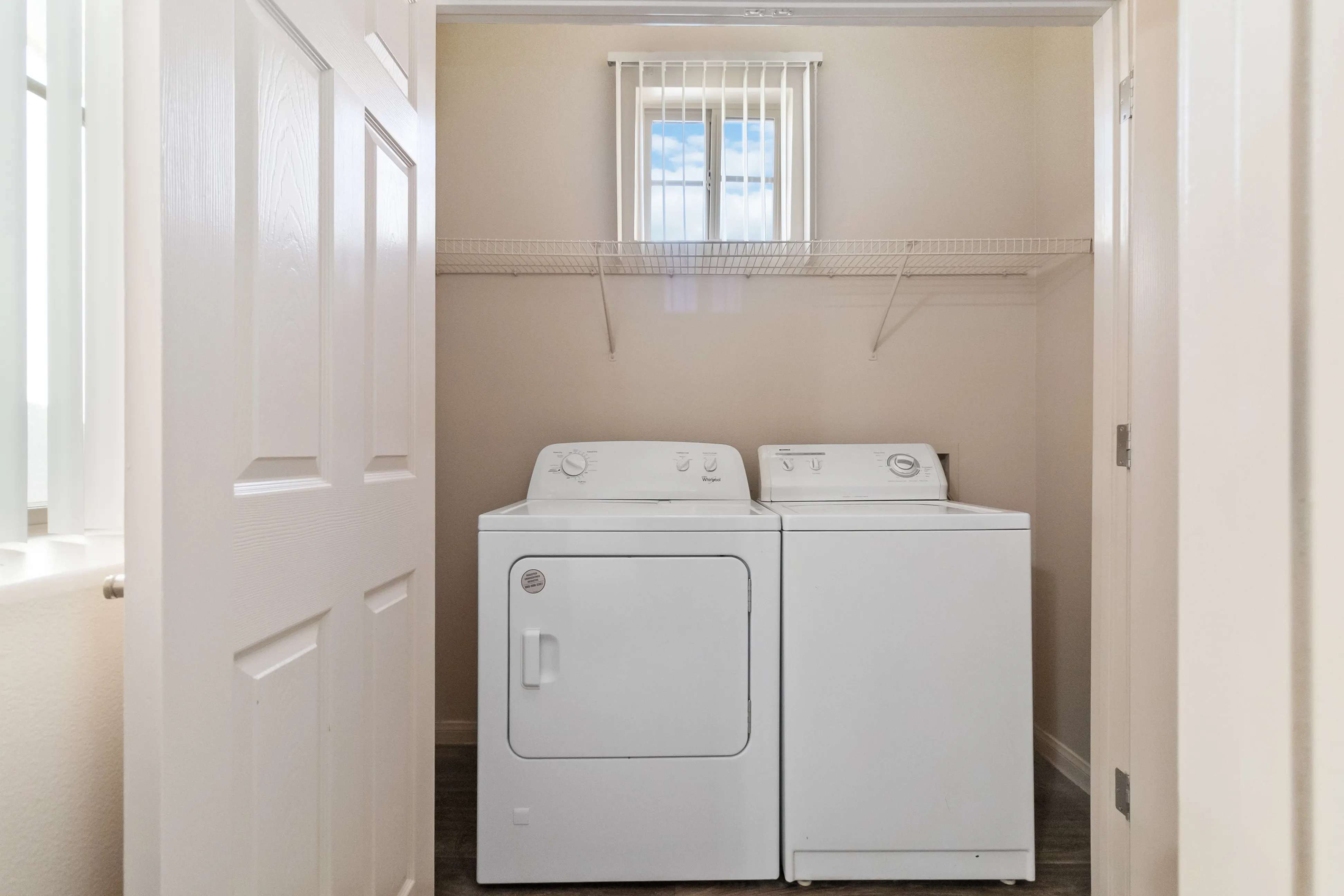Compact laundry room featuring a white washer and dryer set with upper shelving and a window with blinds.