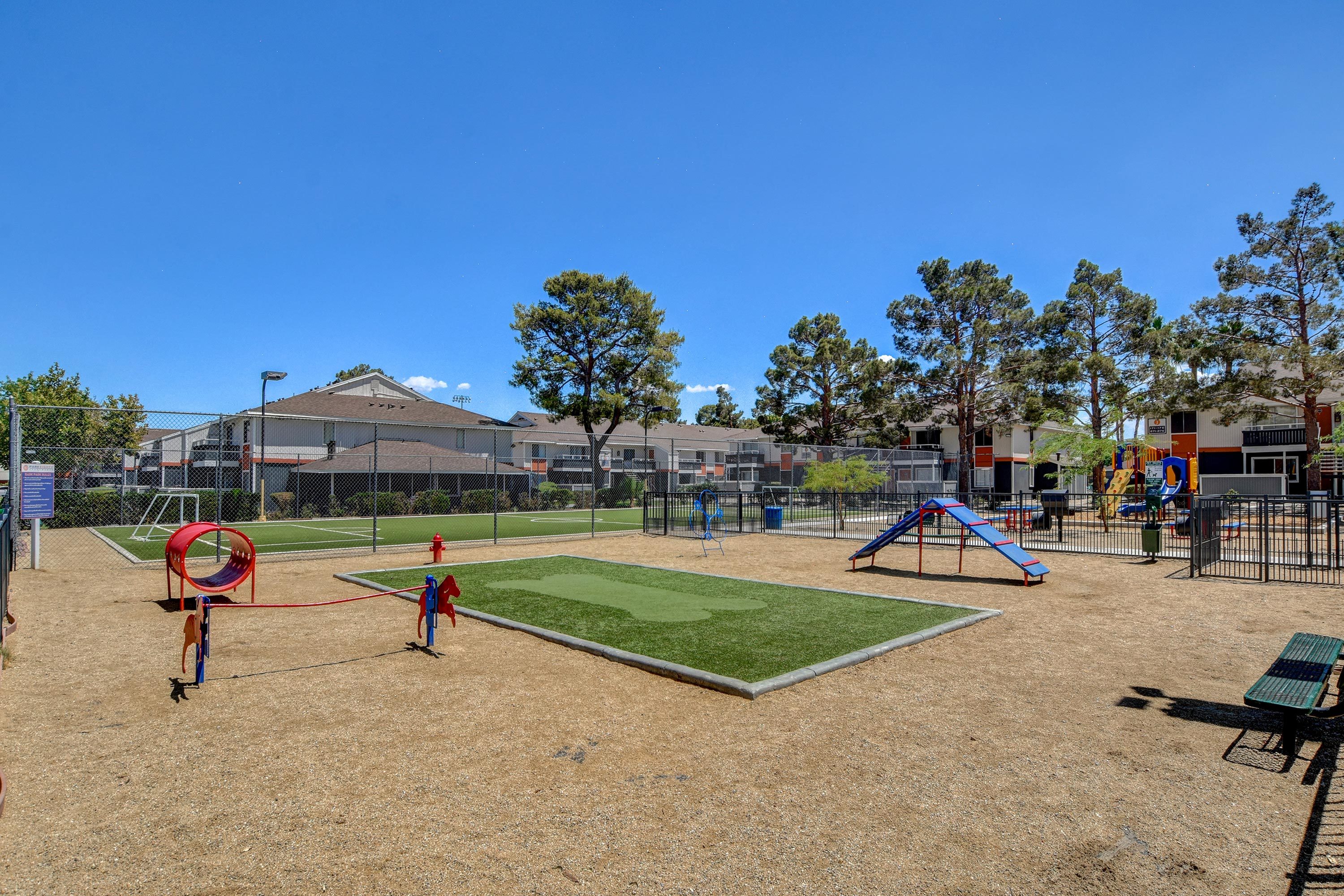 Vibrant community playground featuring colorful slides, climbing structures, and a lush green playfield, set against a backdrop of charming residential buildings and clear blue skies.
