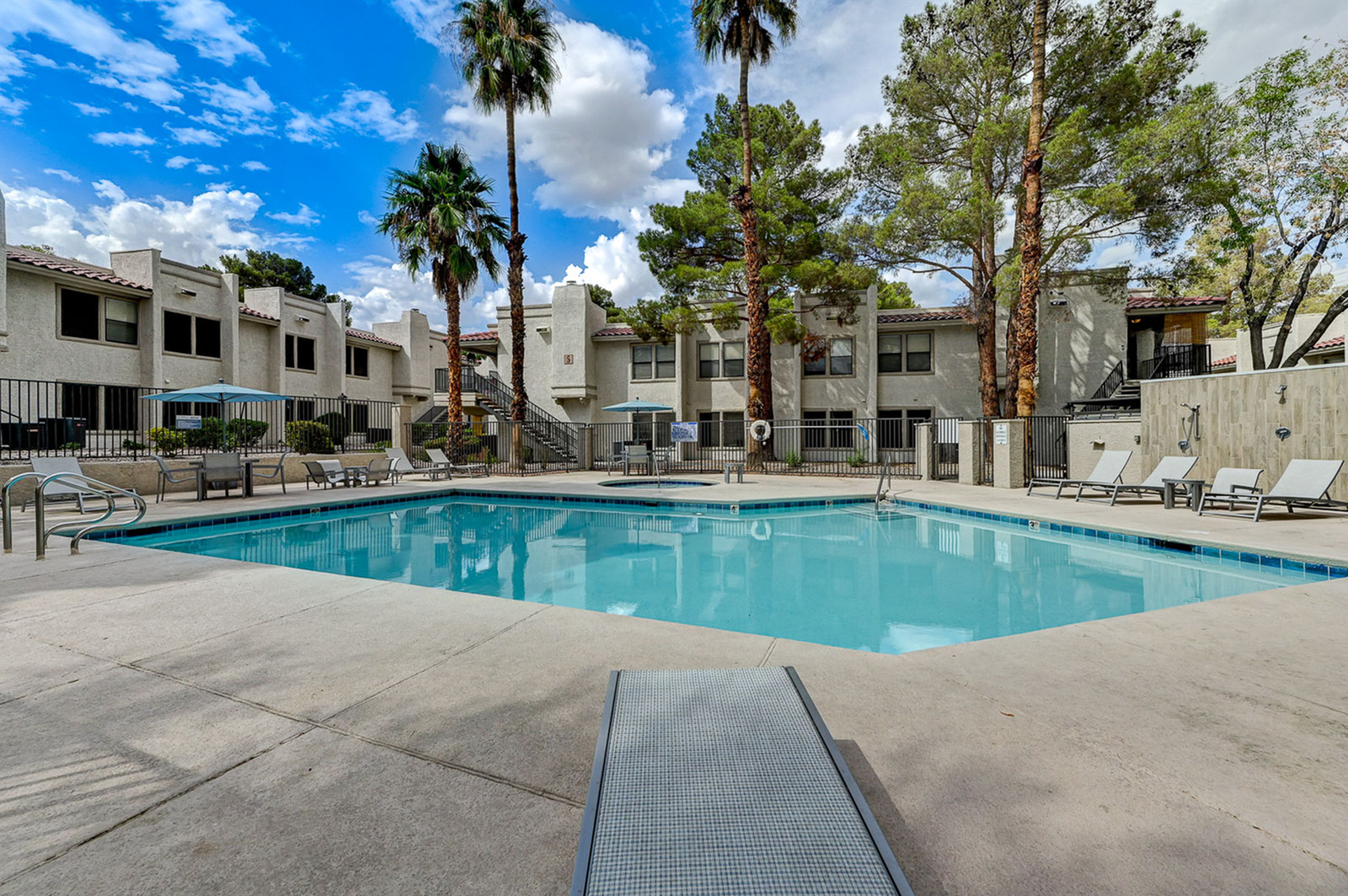 Inviting apartment community featuring a sparkling swimming pool surrounded by palm trees and modern lounge chairs under a vibrant blue sky.