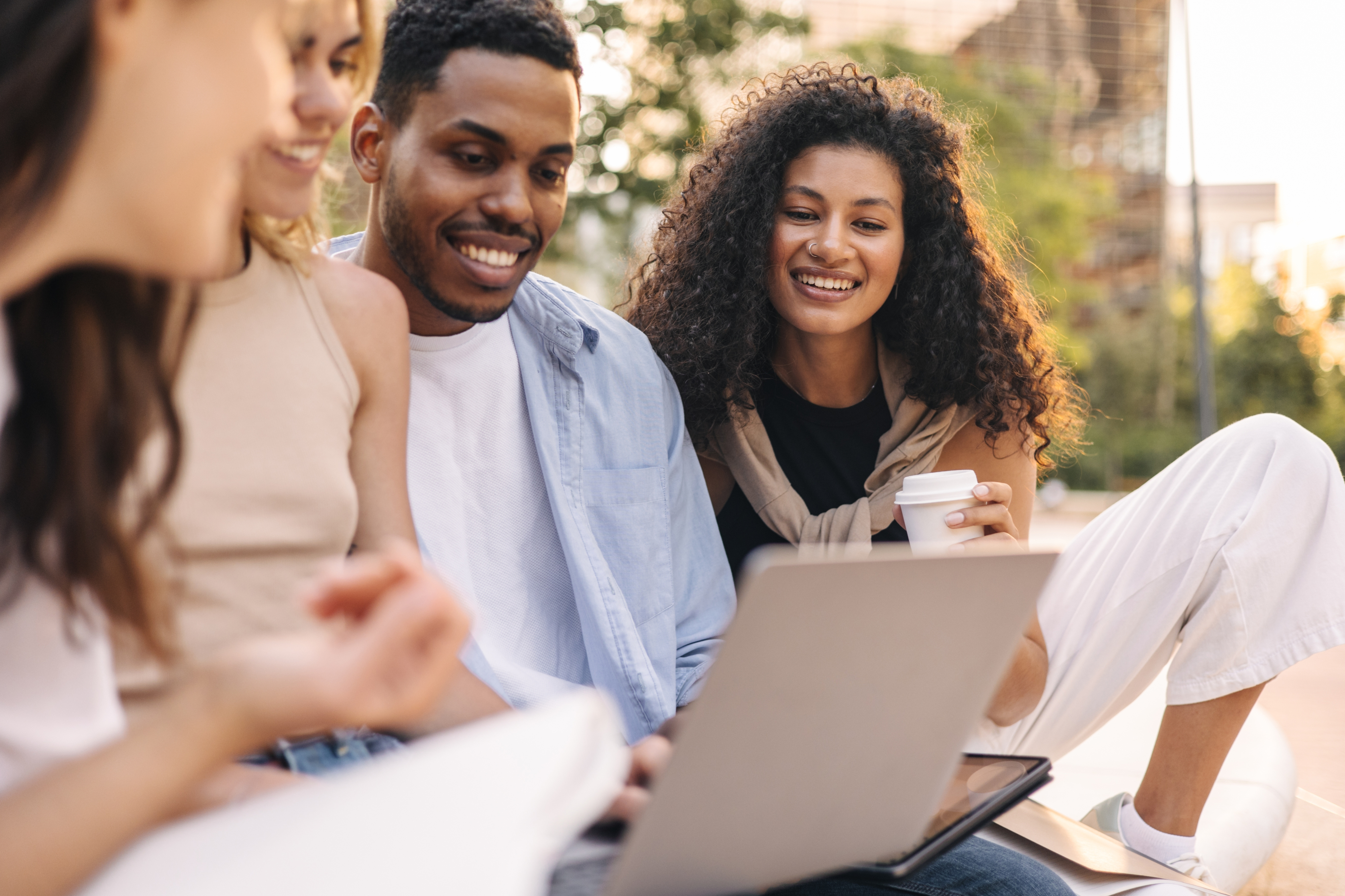 Group of smiling people enjoying an outdoor gathering, engaging over a laptop and coffee.