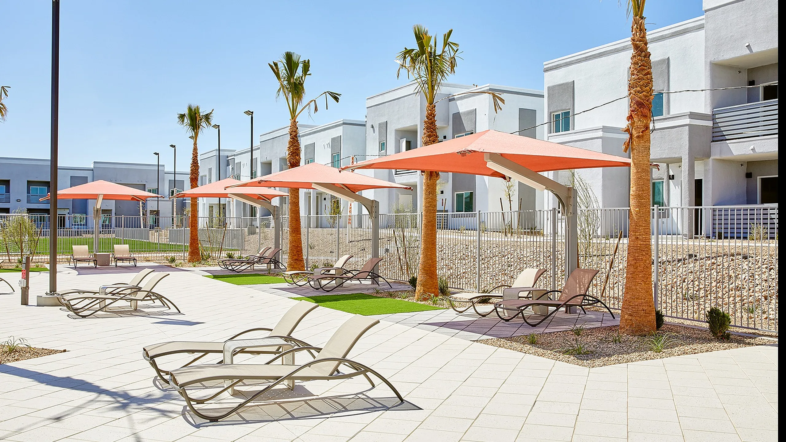 Relaxing outdoor area featuring modern lounge chairs and sunshades, surrounded by tropical palm trees at a contemporary apartment complex.