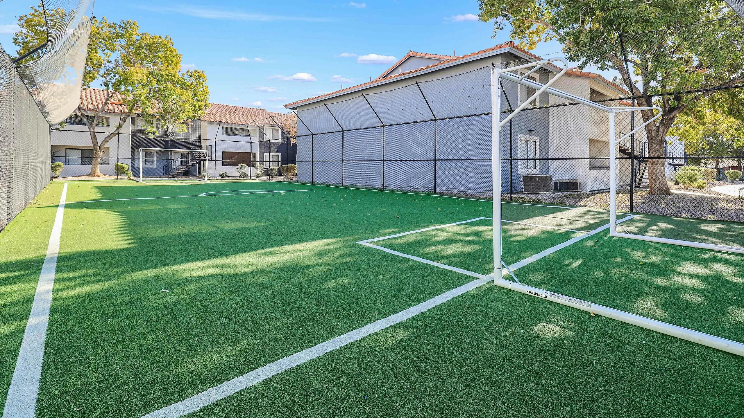 Outdoor soccer field with lush green turf, surrounded by trees and residential buildings on a bright sunny day.