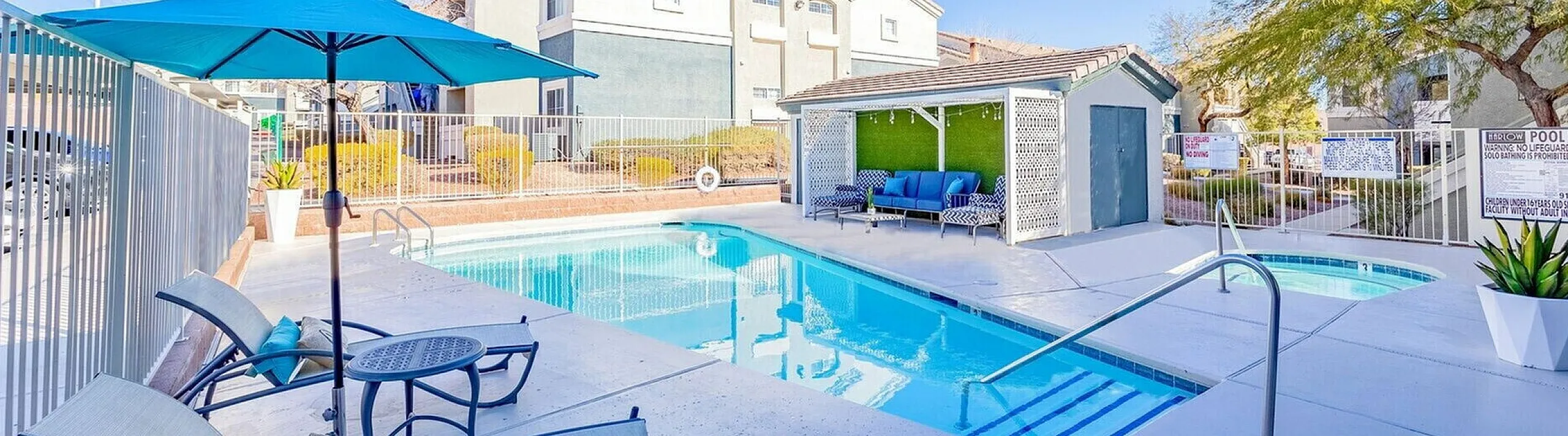 Inviting apartment swimming pool area featuring chic lounge chairs, a vibrant blue umbrella, and a stylish sitting area, perfect for relaxation under sunny skies.