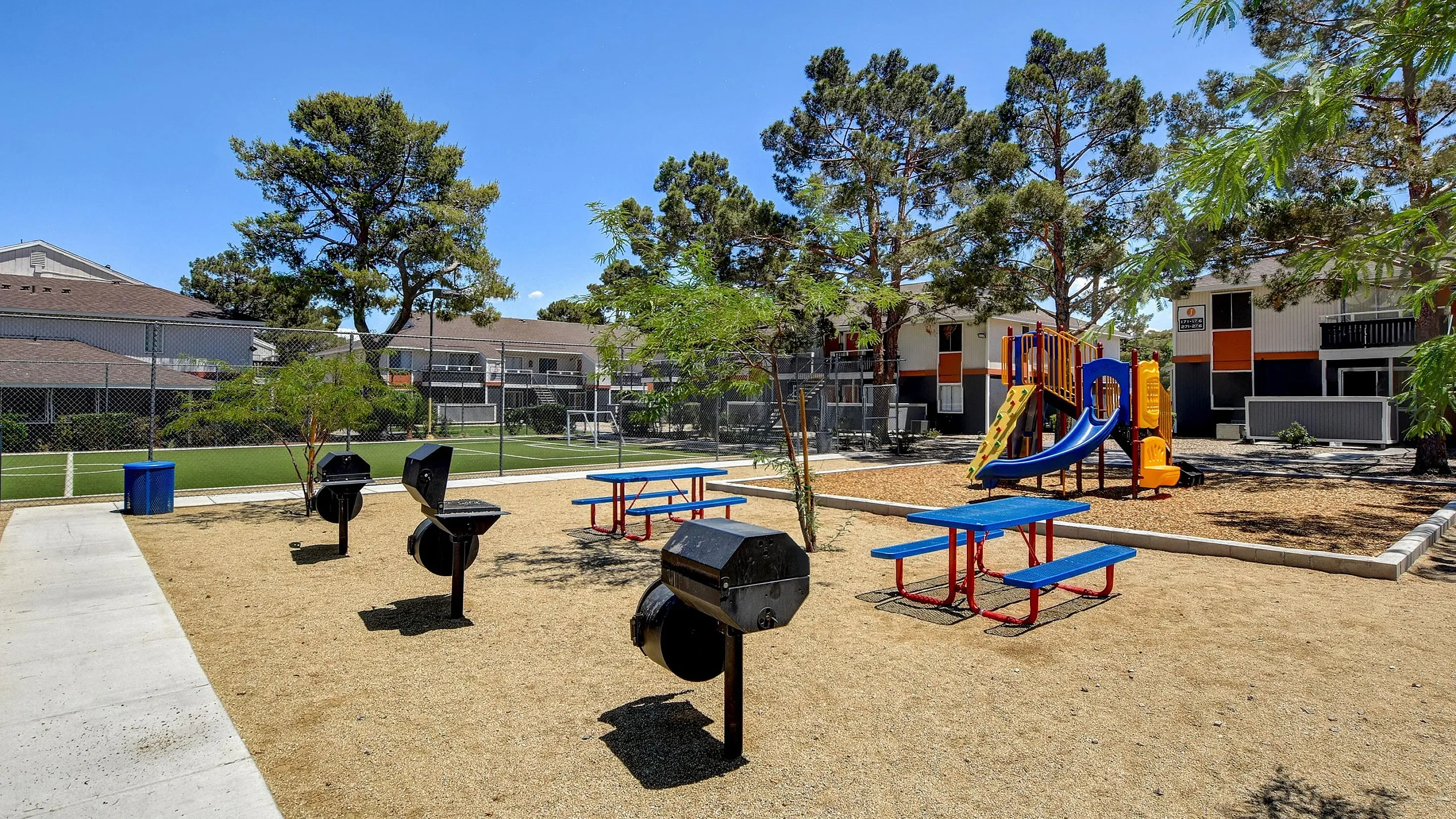 Vibrant community park featuring colorful playground, picnic benches, and barbecue grills, surrounded by lush trees and a soccer field.