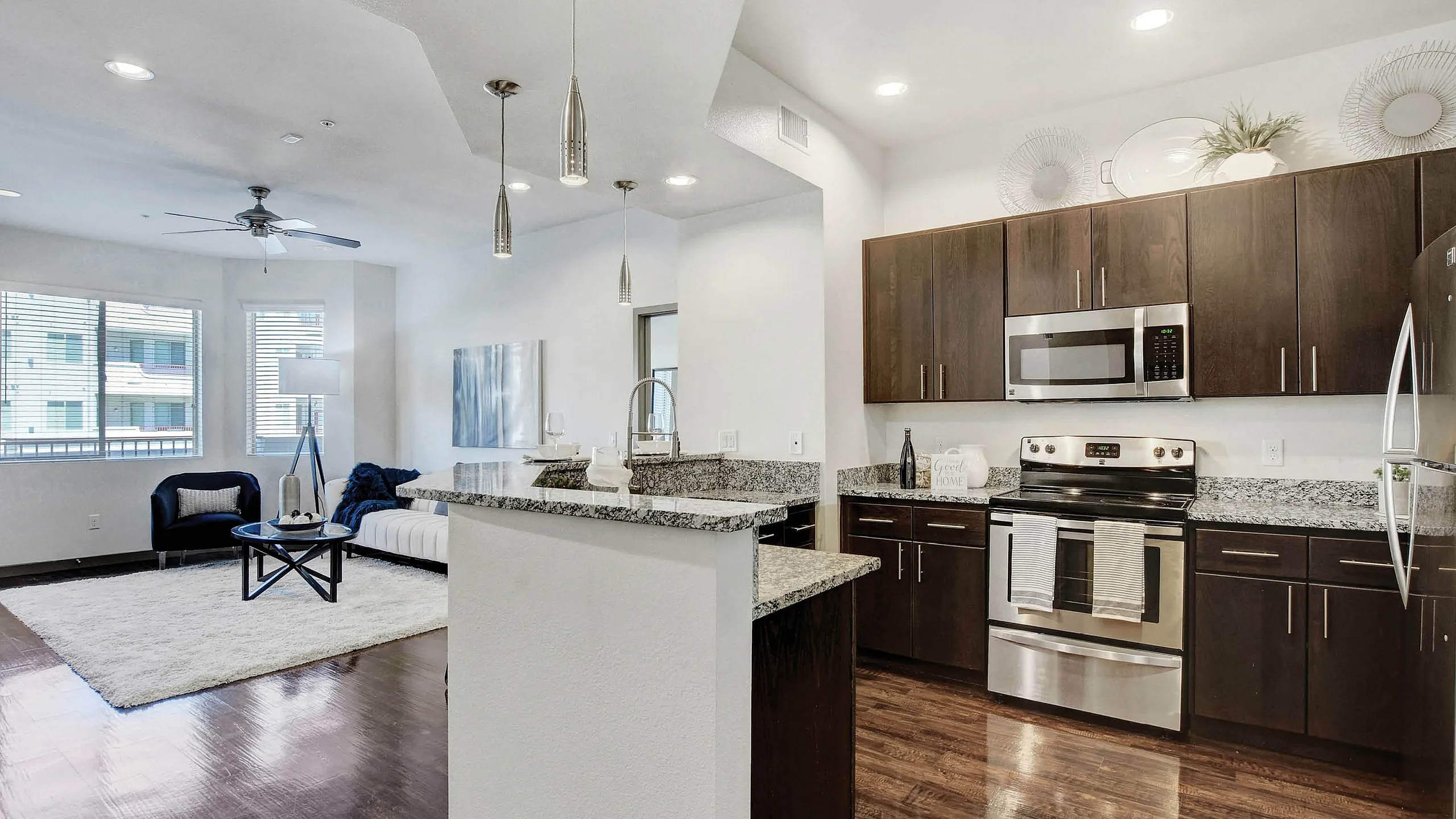 Elegant open-concept living room and kitchen with modern stainless steel appliances, dark wood cabinetry, and chic pendant lighting, highlighted by a cozy seating area and large windows.