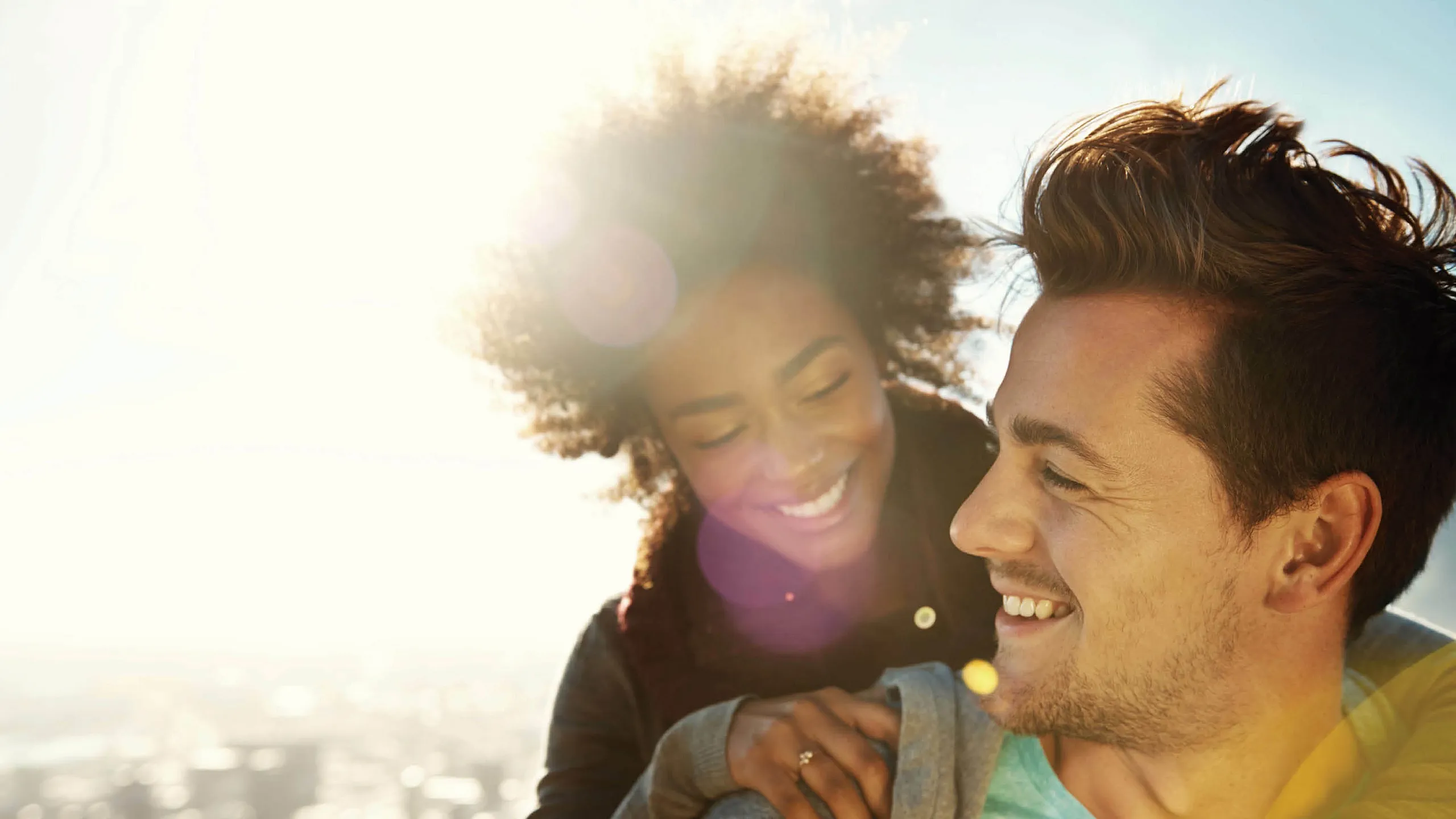 Couple enjoying a sunny day outdoors, smiling and embracing each other with a cityscape in the background.