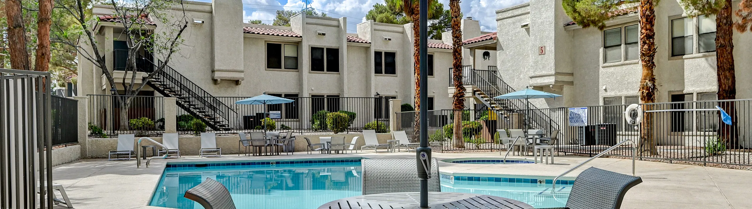 Inviting apartment complex pool area with modern outdoor seating and umbrellas, surrounded by lush trees and stylish buildings under a vibrant blue sky.
