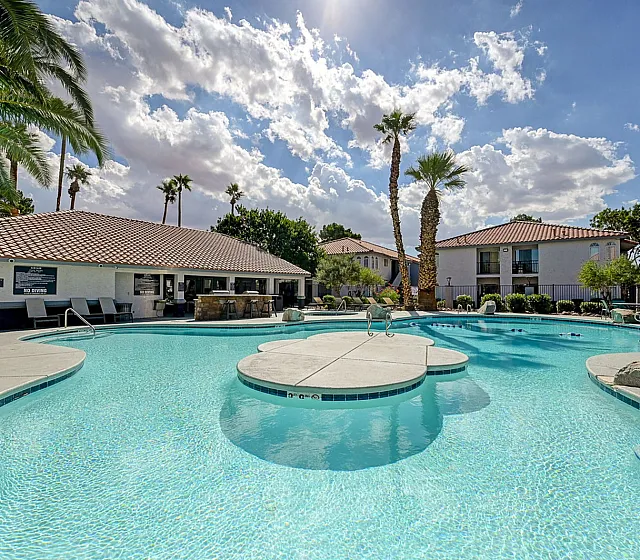 Luxurious apartment complex featuring a stunning outdoor pool, surrounded by palm trees and sun loungers, under a bright blue sky with fluffy clouds.