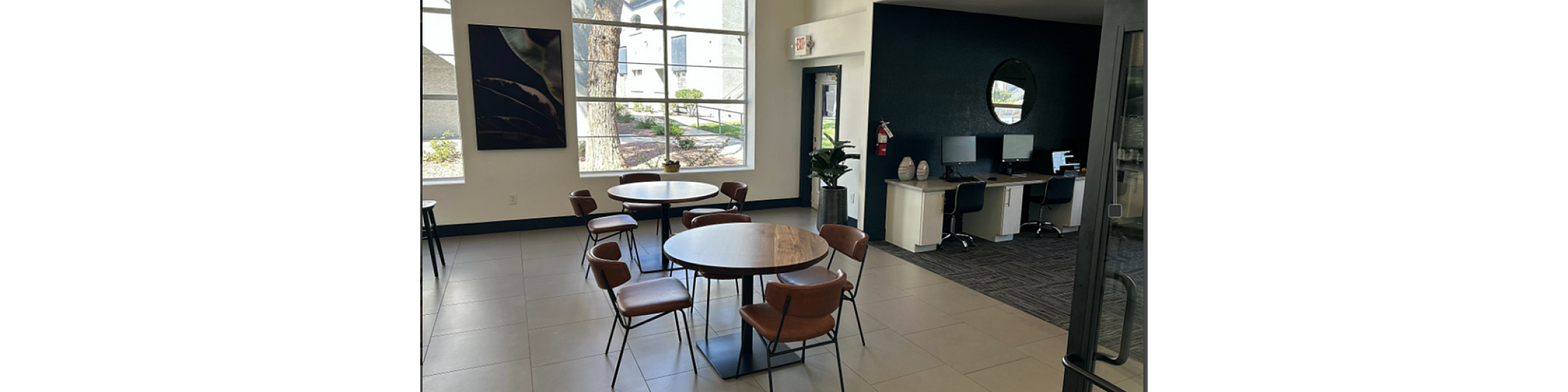 Bright and modern common area with stylish round tables, sleek chairs, and a computer work station in a beautifully lit space.