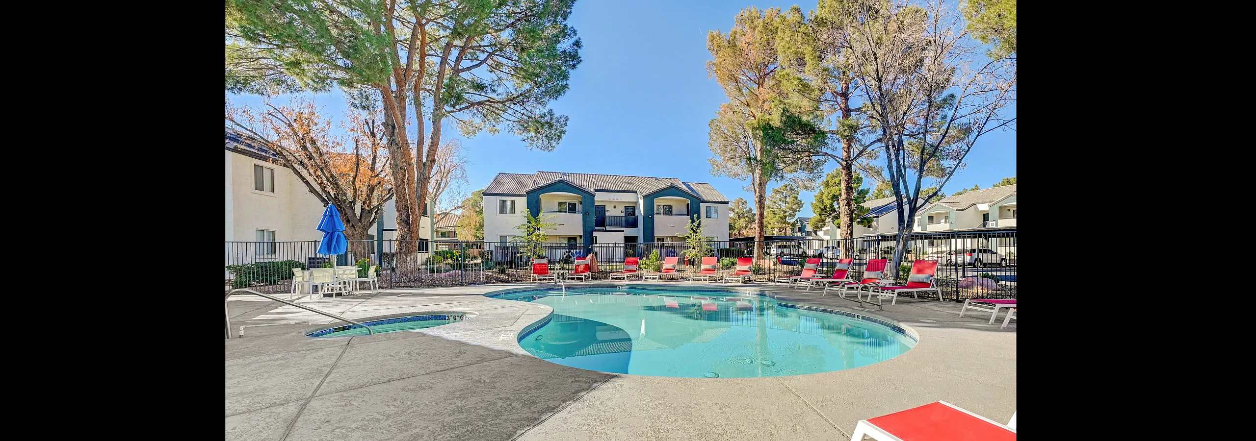 Inviting apartment complex with a sunlit pool area, surrounded by red loungers and shaded by tall trees.