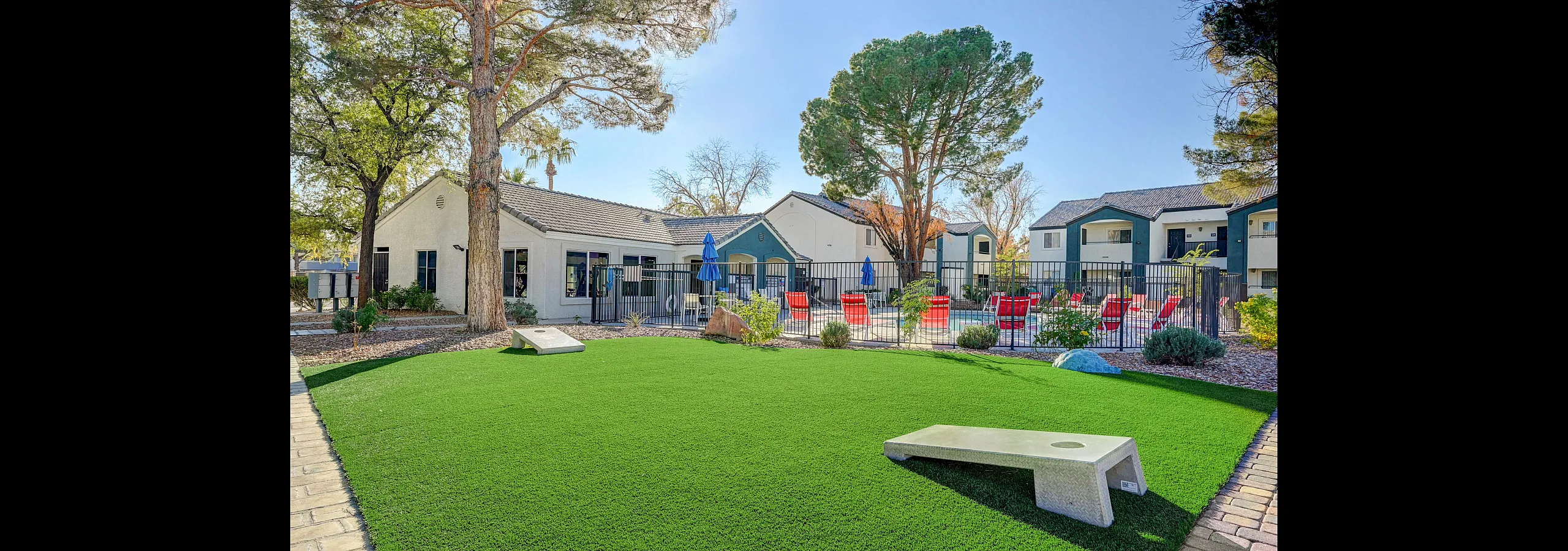 Charming outdoor area with lush green lawn, mature trees, and a refreshing pool surrounded by vibrant red lounge chairs, perfect for relaxation and social gatherings.