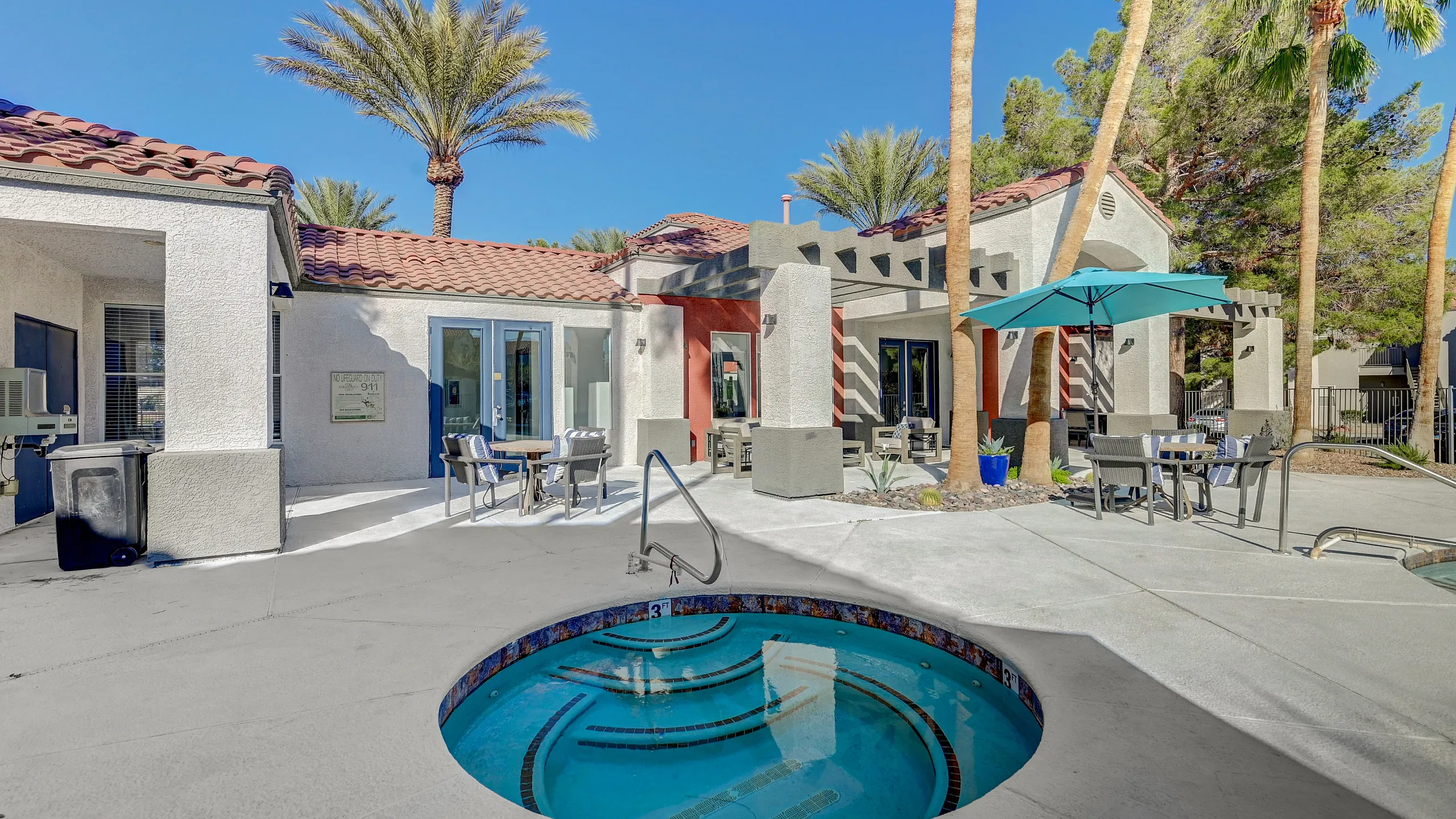 Contemporary outdoor lounge area featuring a luxurious round hot tub, patio seating, and vibrant palm trees under a clear blue sky.