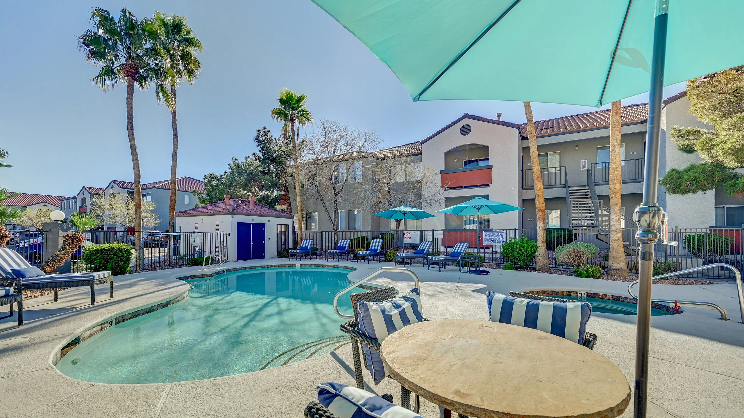 Luxurious apartment complex featuring a serene poolside retreat with stylish loungers, vibrant blue umbrellas, and majestic palm trees under a clear sky.