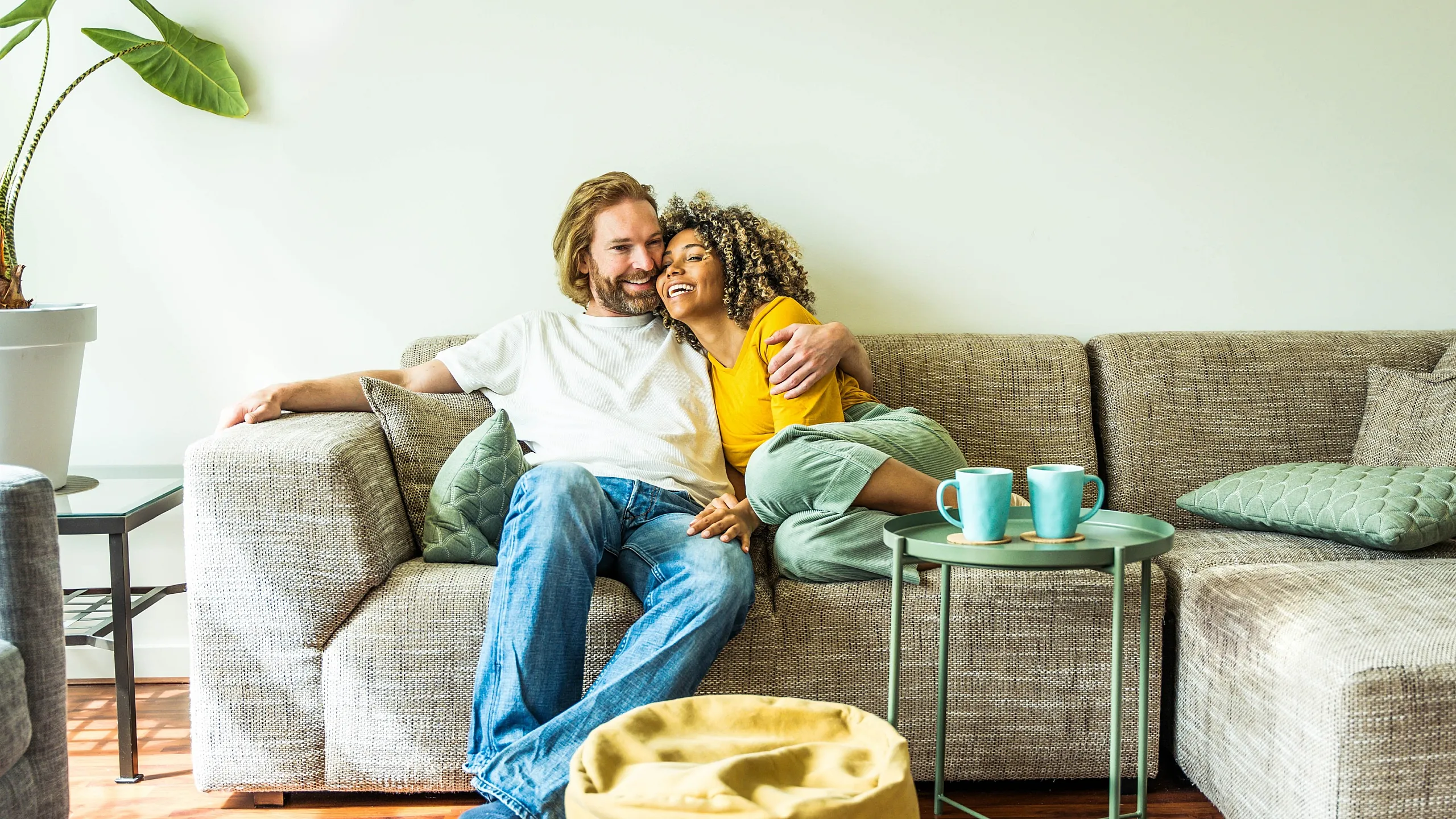 Cozy living room with a comfortable beige sectional sofa, accented by vibrant turquoise mugs and green cushions, perfect for relaxation. Hardwood floors and lush plant add a touch of elegance.