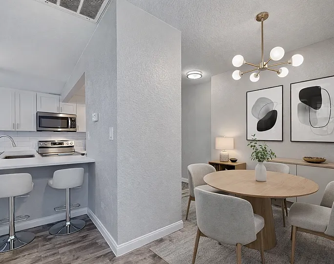 Modern open-plan kitchen and dining area with elegant bar stools, sleek appliances, and contemporary artwork on soft gray walls, illuminated by a chic pendant light.