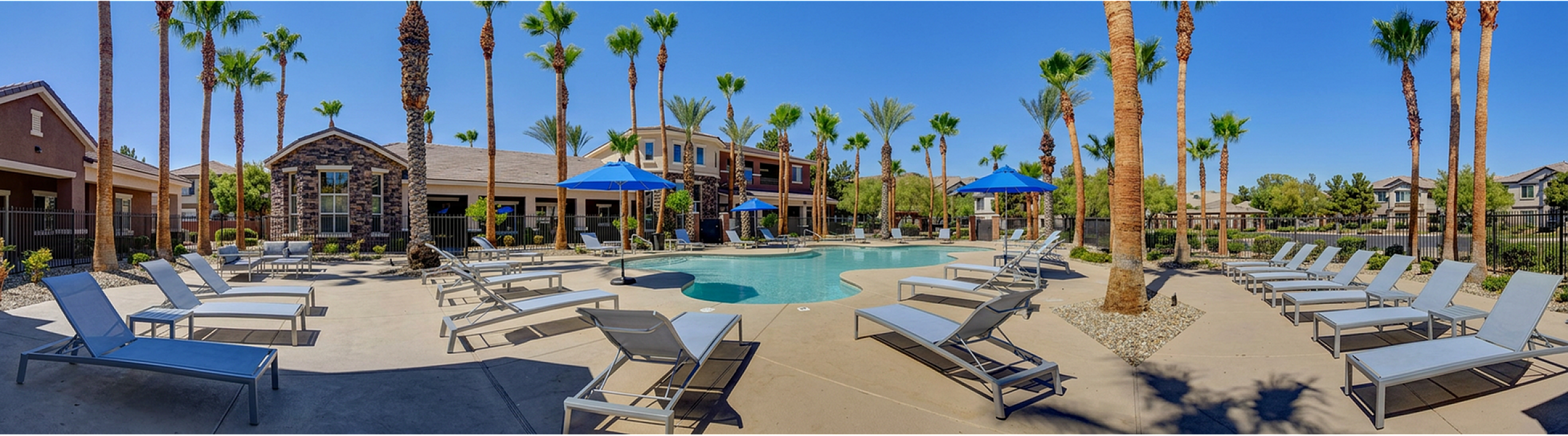 Luxurious apartment complex pool area featuring modern lounge chairs, palm trees, and bright blue umbrellas under a clear sky.