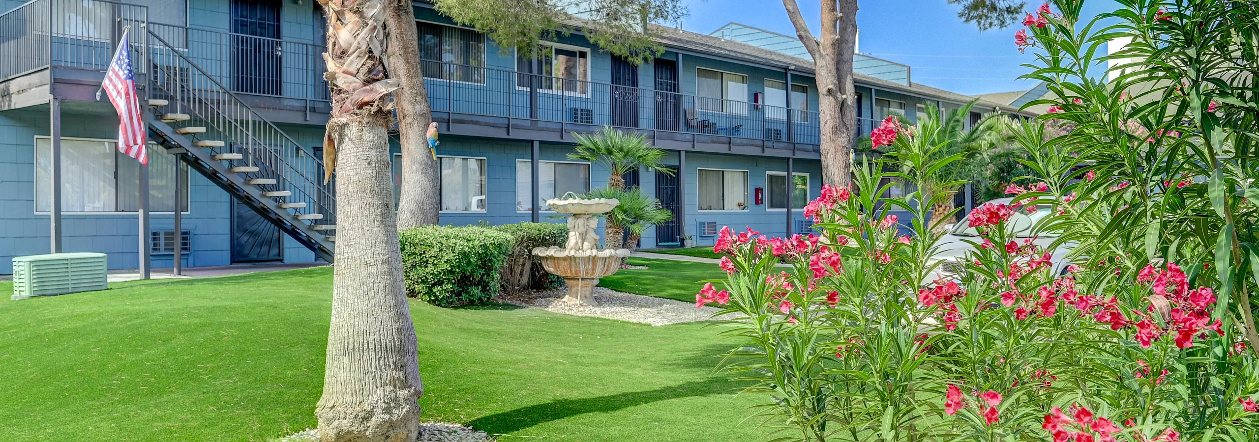 Charming apartment complex with vibrant landscape, featuring lush greenery, blooming flowers, and a serene fountain.