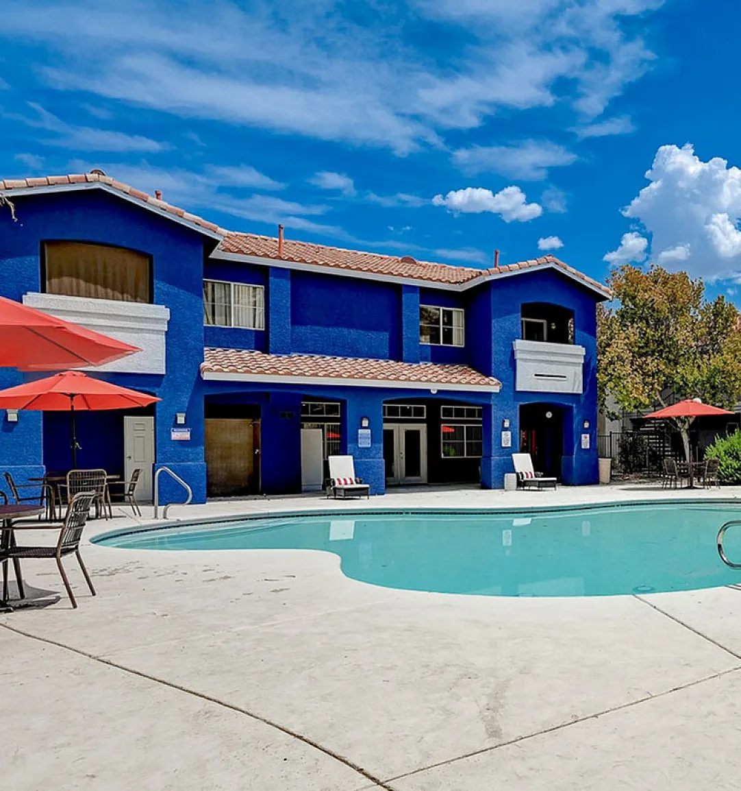 Vibrant apartment complex with a sparkling pool, modern blue accents, and inviting outdoor seating under red umbrellas.