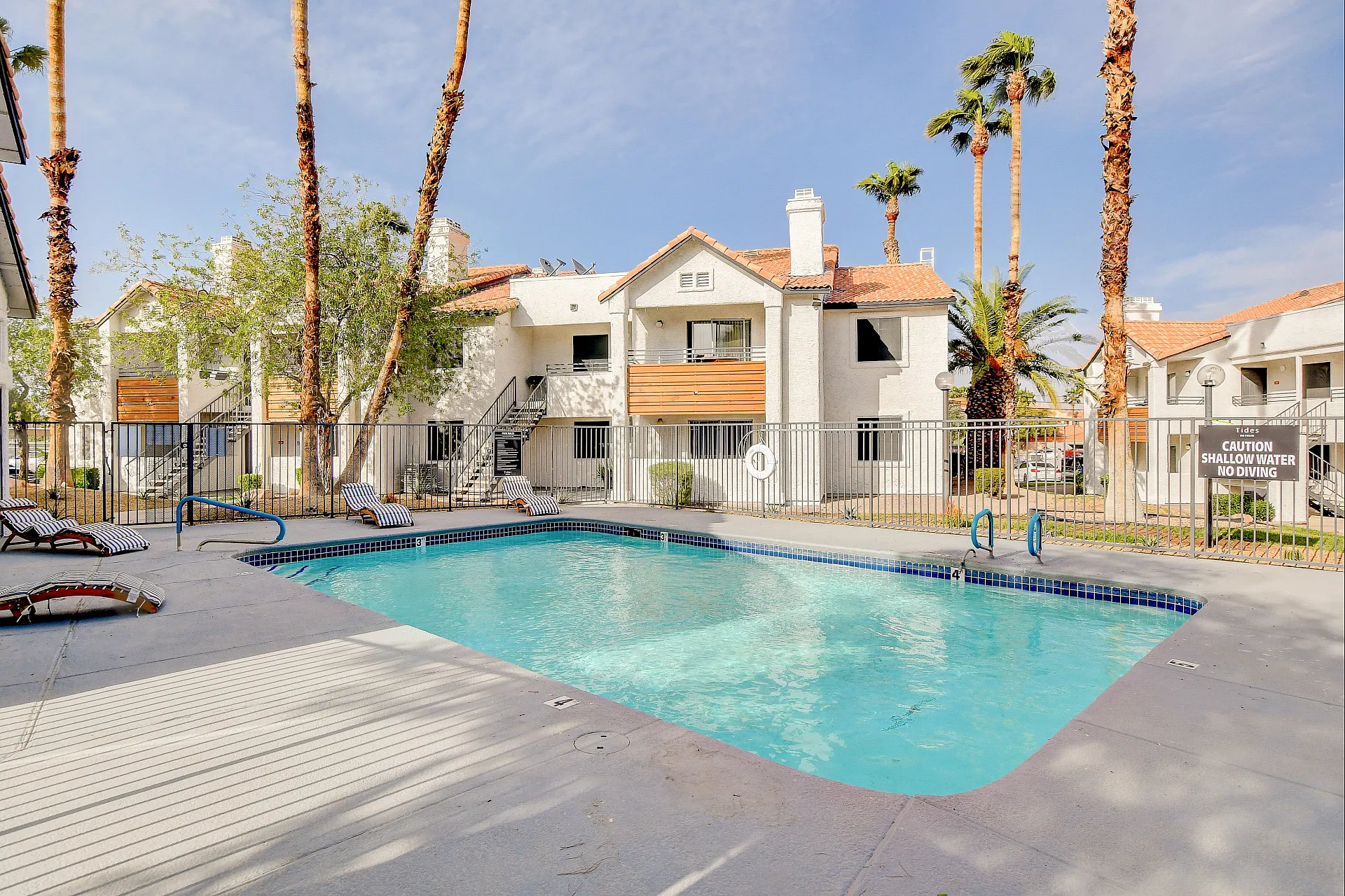 Charming apartment complex with a sparkling outdoor swimming pool surrounded by lush palm trees and inviting lounge chairs.