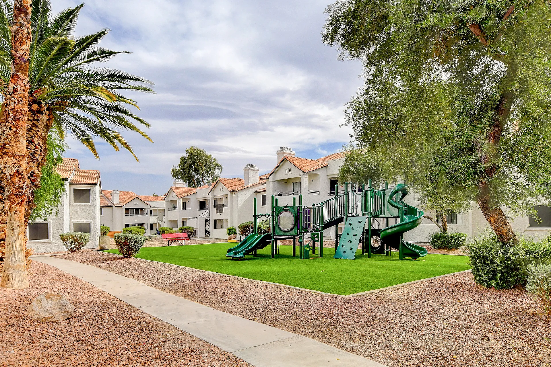 Inviting apartment complex with a lush green playground, surrounded by charming red-roofed buildings and palm trees, offering family-friendly living.