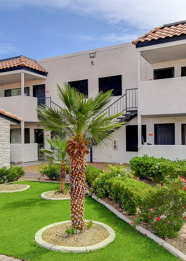 Charming apartment exterior with modern white facade, terra-cotta roof, and lush landscaped gardens featuring palm trees and vibrant greenery.