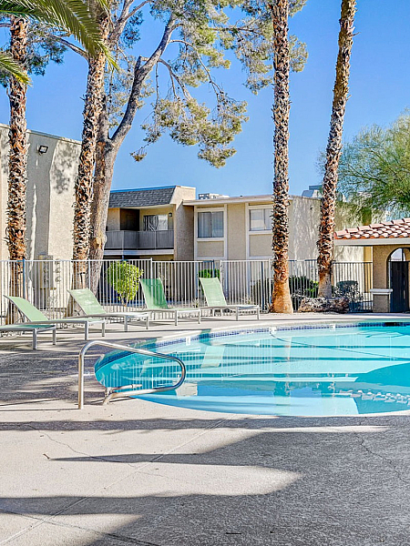 Inviting apartment complex pool area with palm trees, lounge chairs, and a shaded seating area under a blue sky.