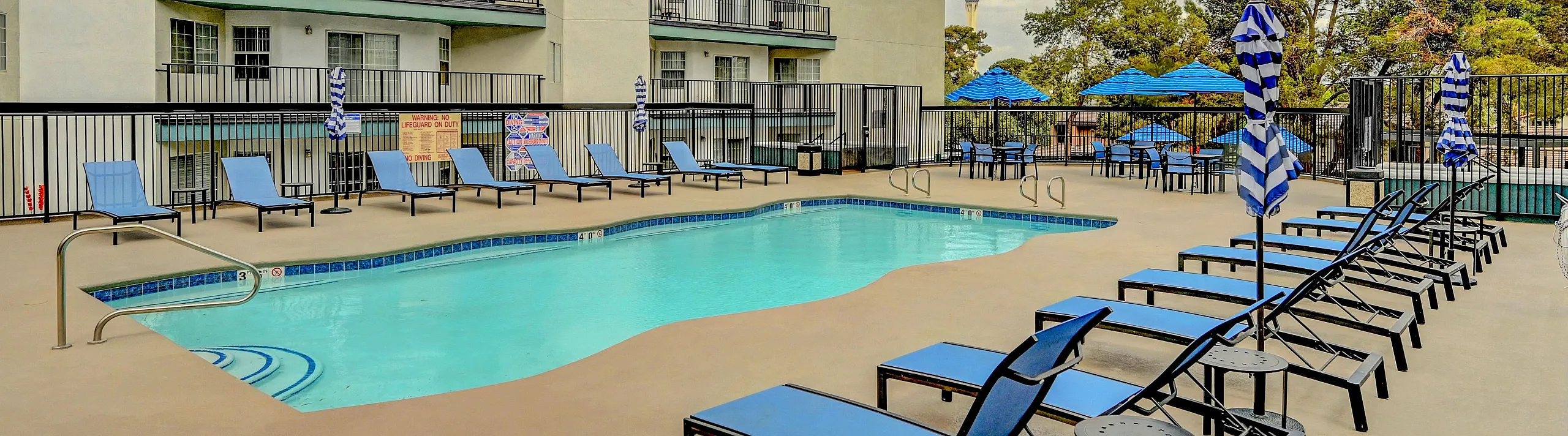 Inviting apartment complex with a pristine outdoor pool, surrounded by modern lounge chairs and vibrant umbrellas, perfect for relaxation.
