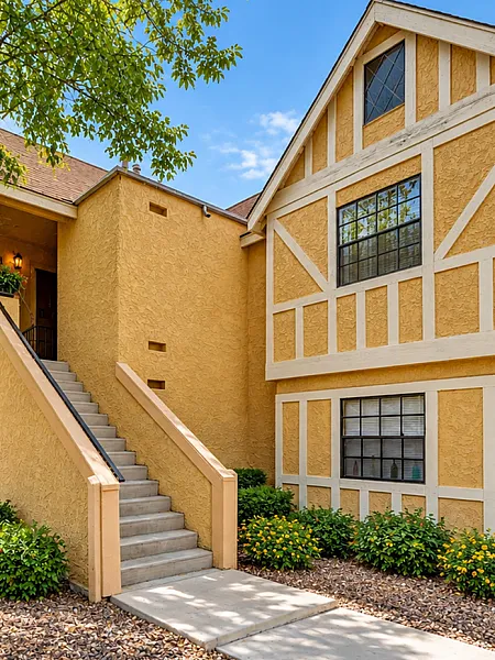 Charming yellow-tan apartment exterior with welcoming stairs, lush greenery, and large windows under a bright blue sky.