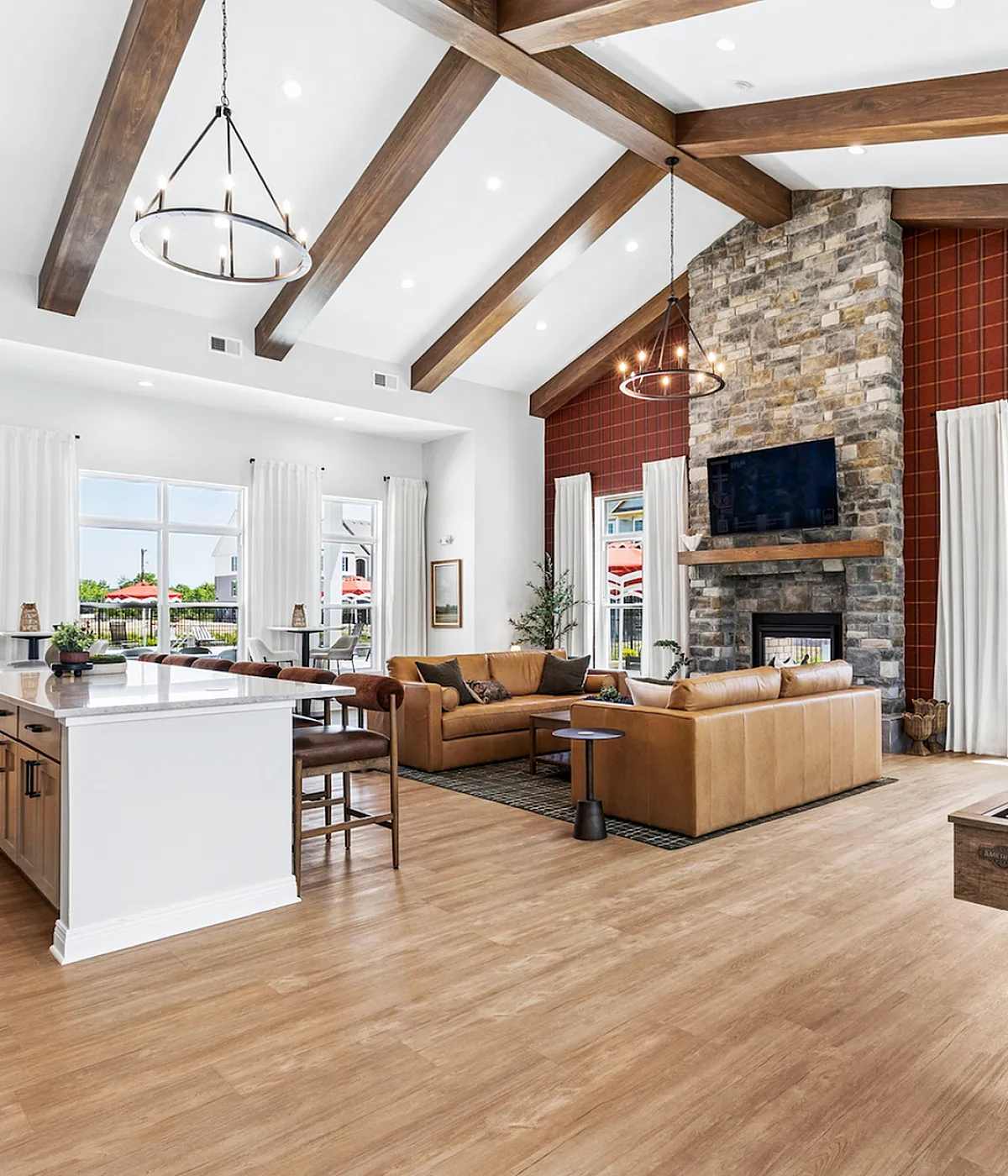 Inviting open-concept living room with vaulted ceilings and elegant wooden beams, featuring a cozy stone fireplace, stylish leather couches, and a sleek shuffleboard table, basking in natural light from large windows.
