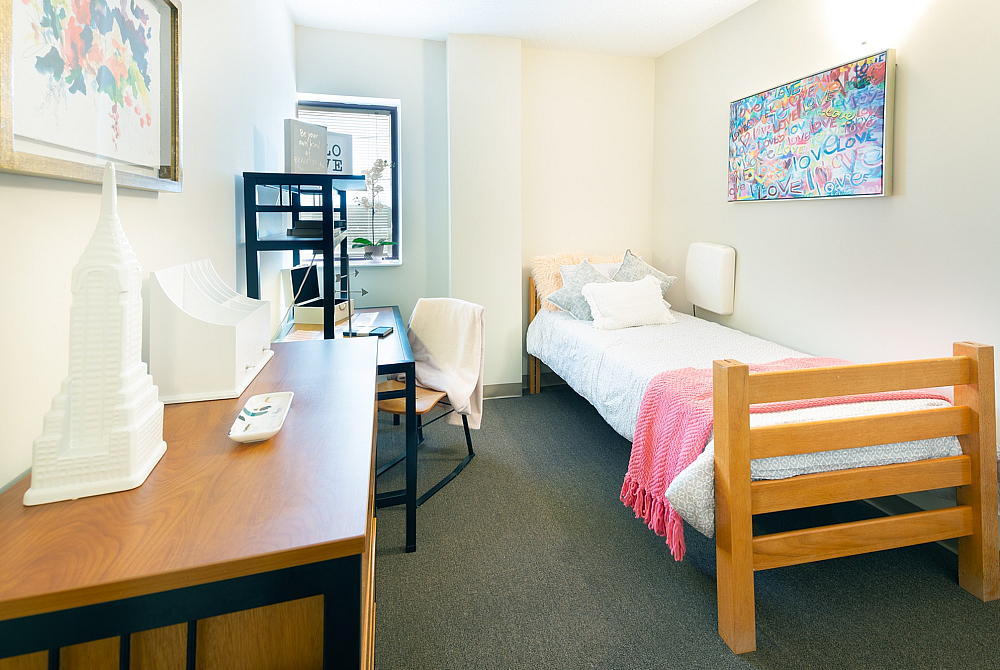 Model apartment bedroom with bed, desk, chair, and dresser in a light wood