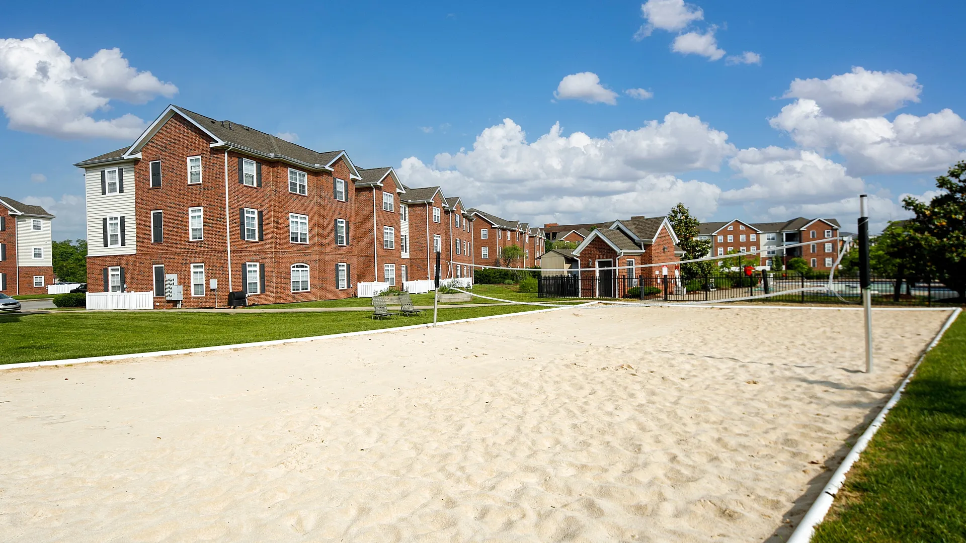 Charming brick apartment complex with lush green lawns and a pristine sand volleyball court under a bright blue sky.