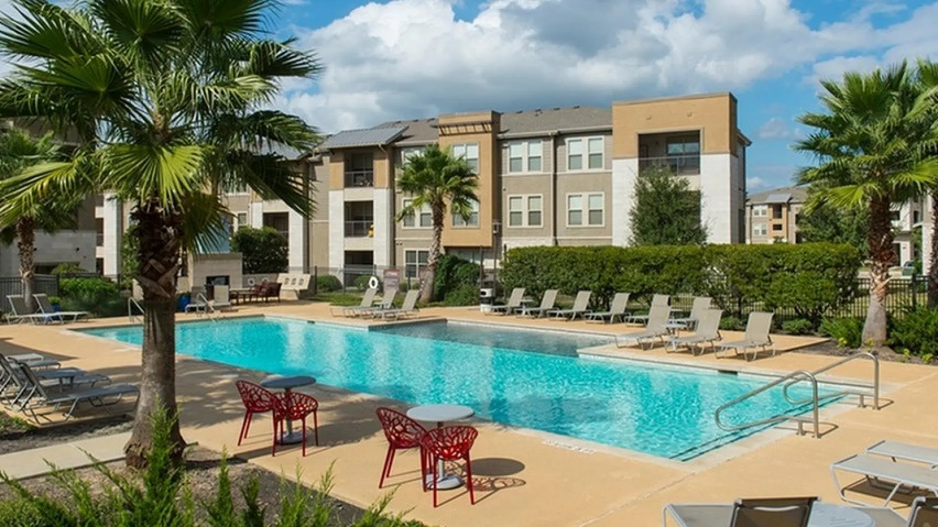 Chic apartment complex with a sparkling outdoor pool, surrounded by lush palm trees and comfortable lounge chairs, under a bright blue sky.