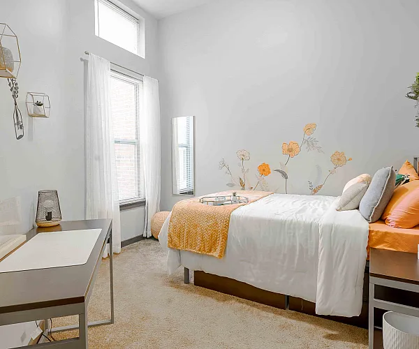 Charming bedroom with a cozy bed adorned with orange and white linens, decorative floral wall art, and modern geometric shelves. Bright natural light filters through the window, enhancing the serene and airy ambiance.
