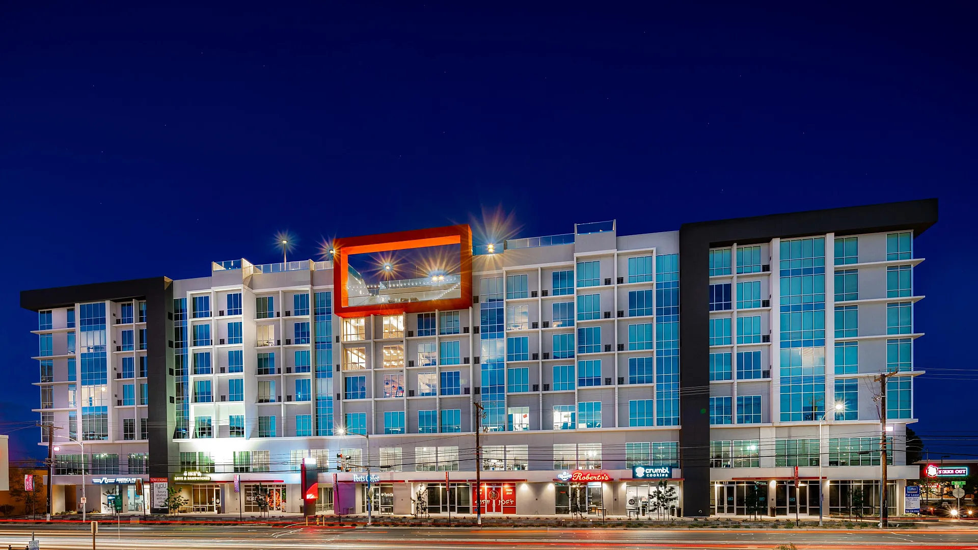 Modern apartment complex at night with vibrant lighting, offering retail shops on ground level and stylish residences above.