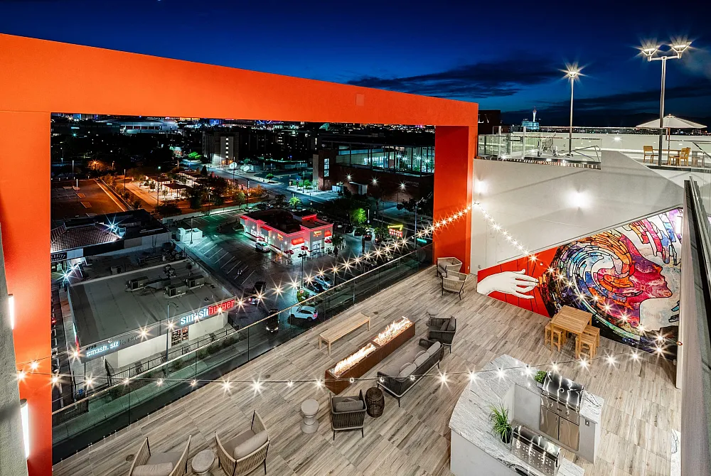 Vibrant rooftop lounge with modern seating, artistic mural, and stunning city views illuminated by string lights.