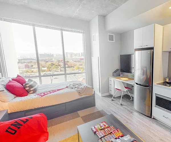 Bright modern bedroom featuring a comfortable bed, large TV with stylish cabinetry, cozy grey armchair, and expansive city views from floor-to-ceiling windows.
