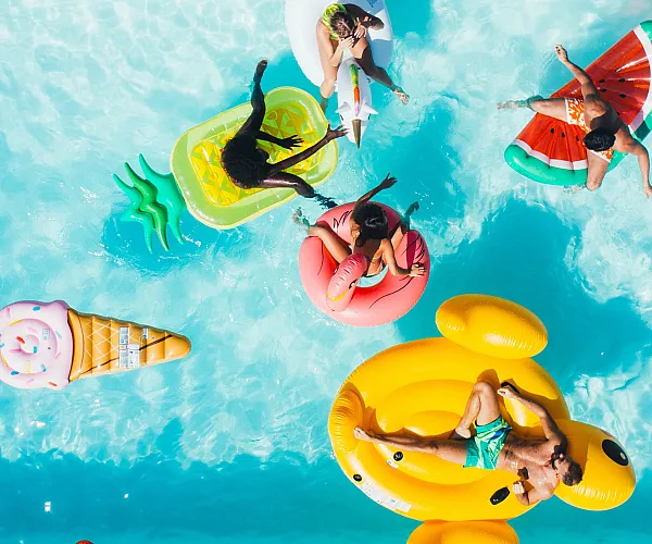 Vibrant aerial view of a swimming pool with people enjoying colorful inflatable floats shaped like a unicorn, pizza, ice cream cone, and watermelon.