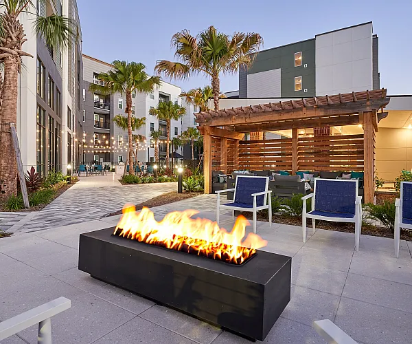 Chic apartment courtyard featuring a modern fire pit, comfortable seating, and ambient string lighting under palm trees.