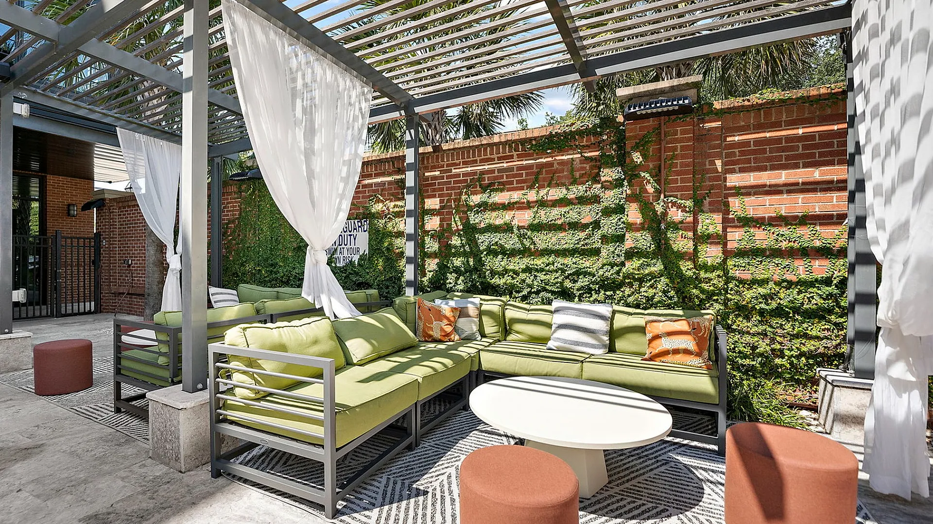 Chic outdoor lounge area with modern pergola, cozy green cushions, lush ivy-covered brick wall, and stylish round table.